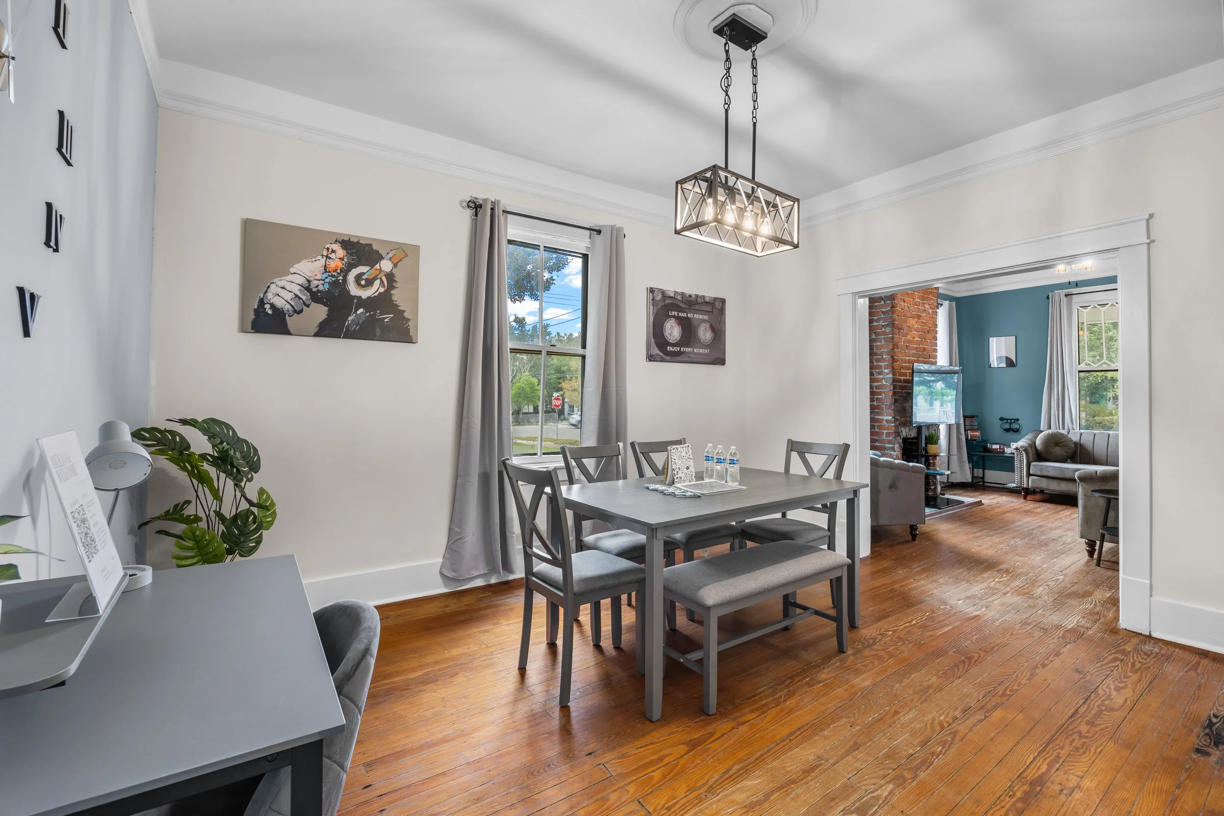 Dining room with gray table and chairs, hardwood floor, artwork on the wall, and adjacent living room with brick wall, sofa, and TV.