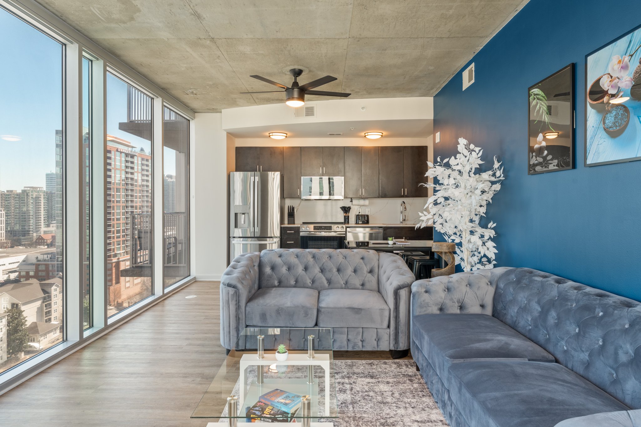 Modern apartment living room with large windows, two gray tufted sofas, a glass coffee table, and a blue accent wall with framed artwork, overlooking an urban cityscape.