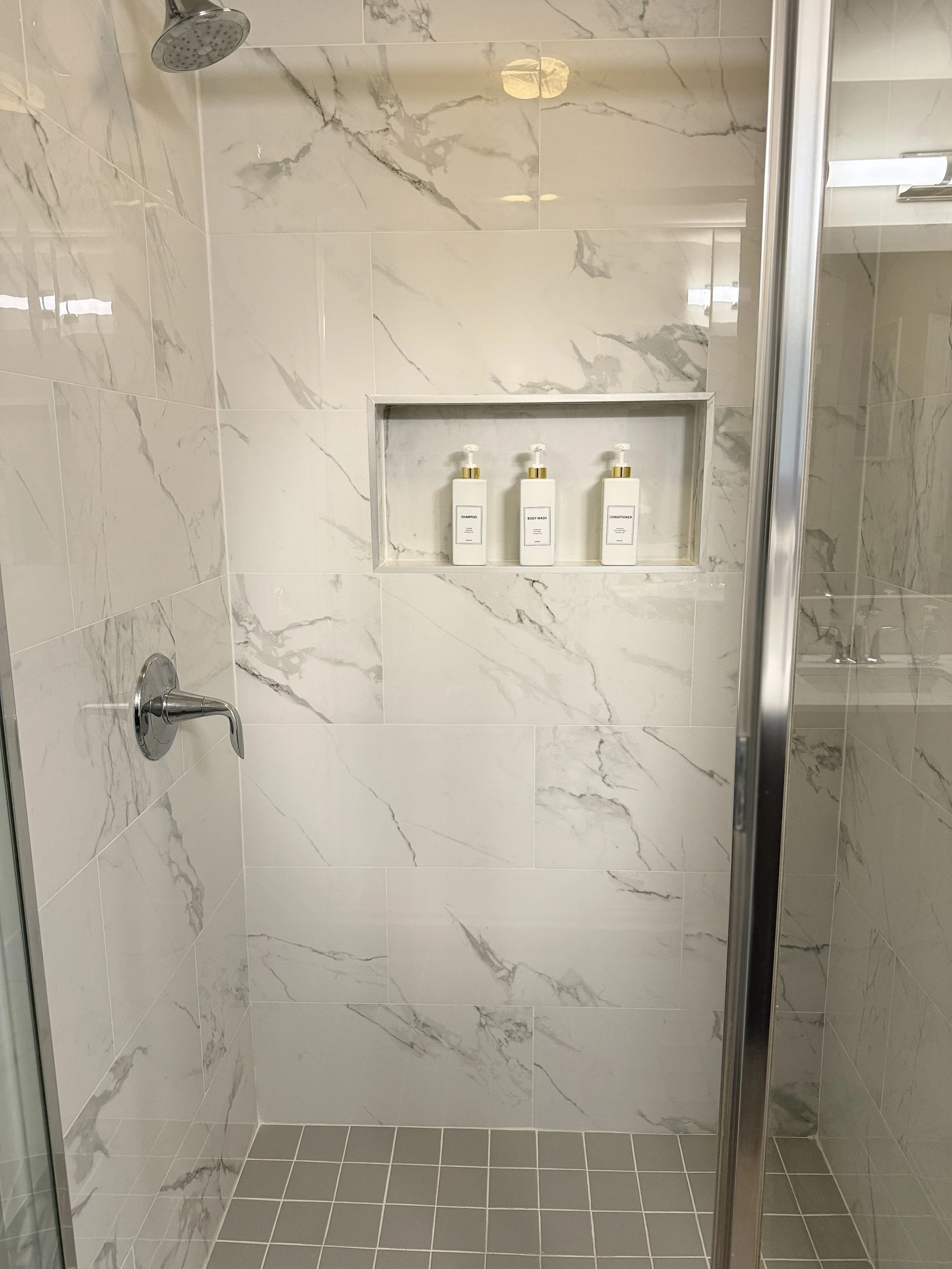 Modern shower with marble tile walls, a rainfall showerhead, and a built-in niche containing three bottles of shampoo, body wash, and conditioner.