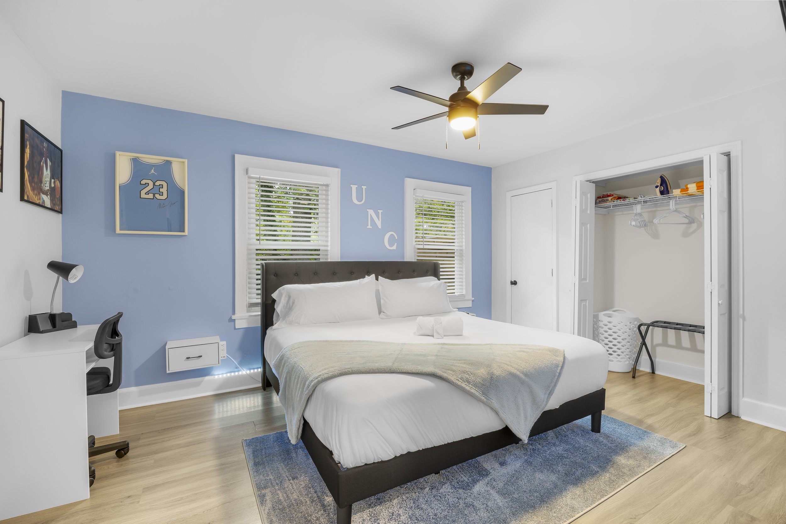A bedroom with a white bed, dark gray headboard, light-colored blanket, and white pillows. There are two windows behind the bed with white blinds, and the wall is painted blue with white letters spelling 'UNC'. A ceiling fan with five blades is overh