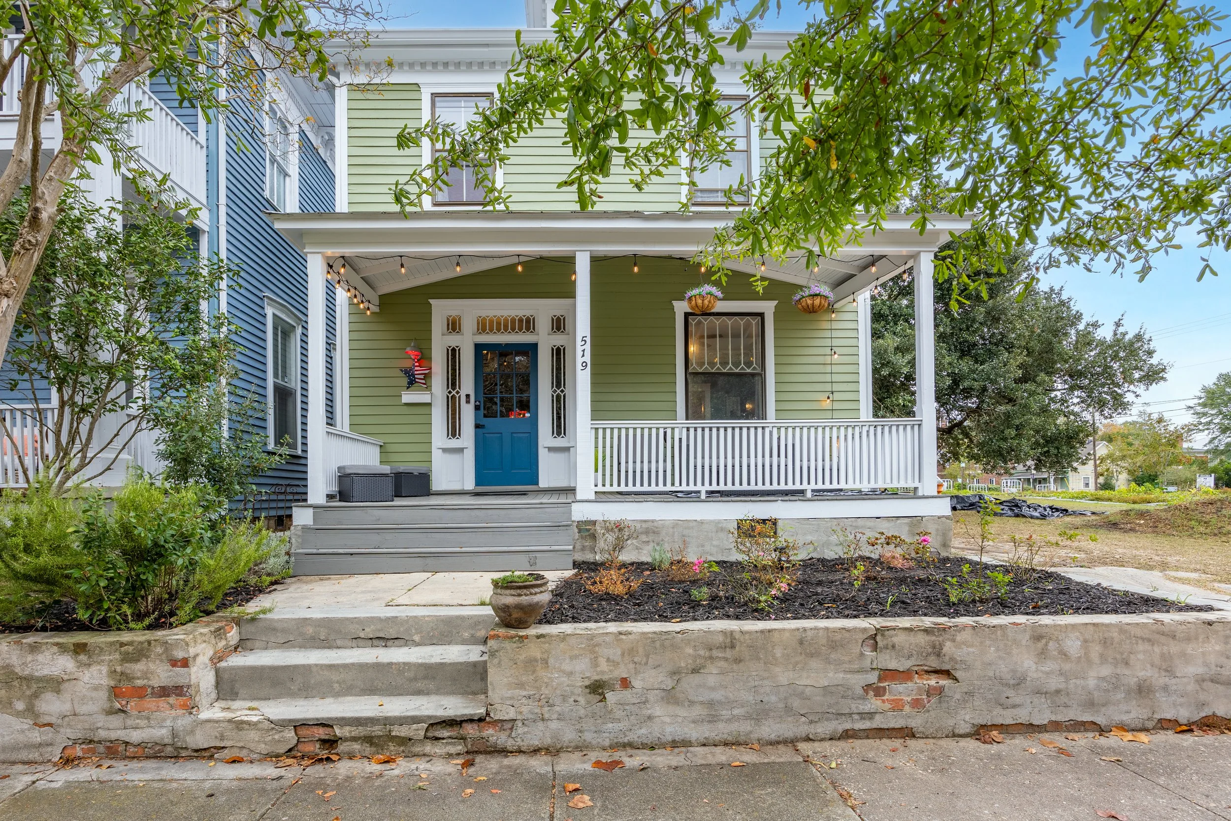 Front porch of a colorful house with a green facade, white trim, and blue door, decorated with hanging plants, string lights, and outdoor furniture, surrounded by a garden with plants and trees.