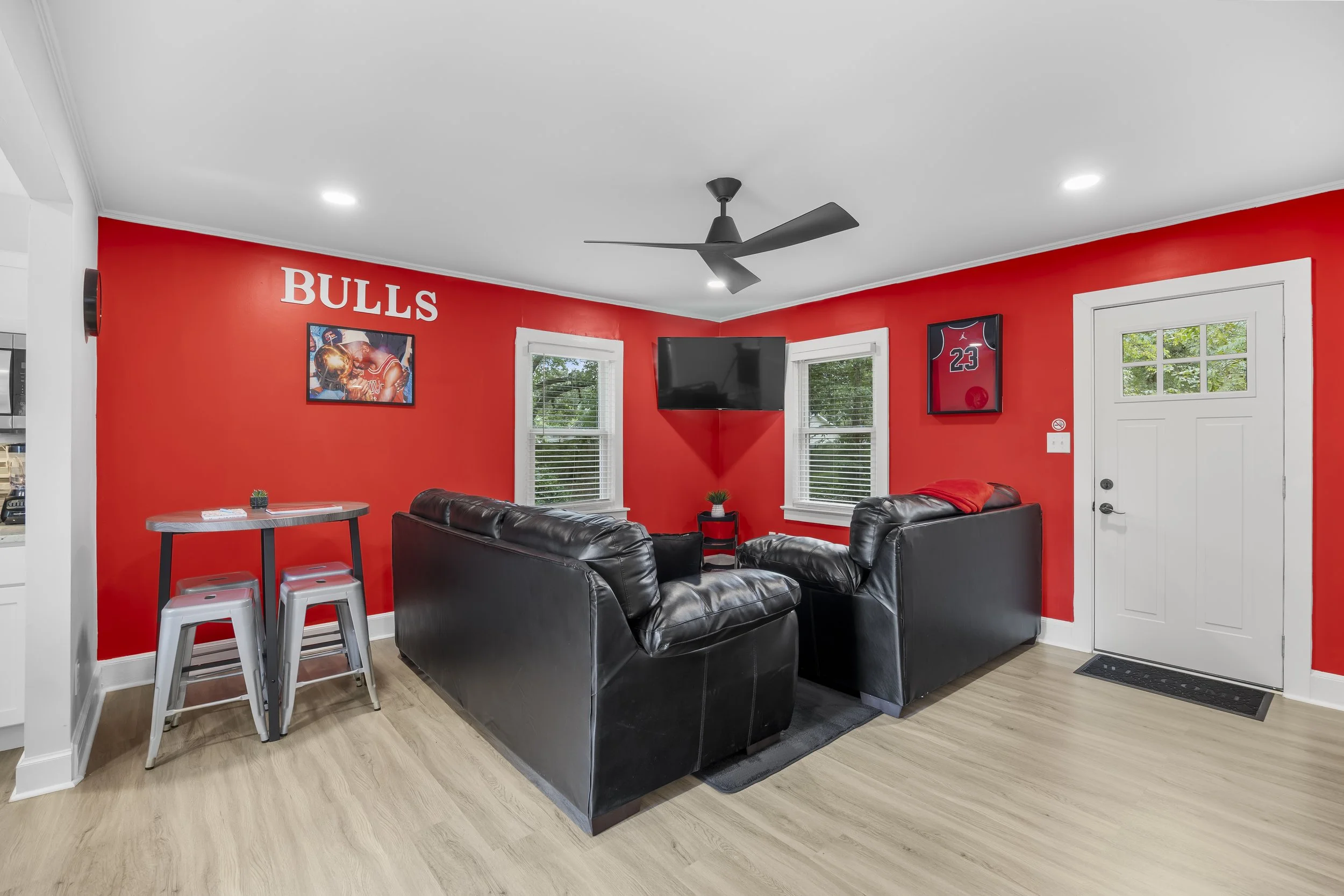 Living room with red walls, black leather sofas, a wall-mounted TV, framed sports memorabilia, a small round table with two stools, two windows, and a ceiling fan.