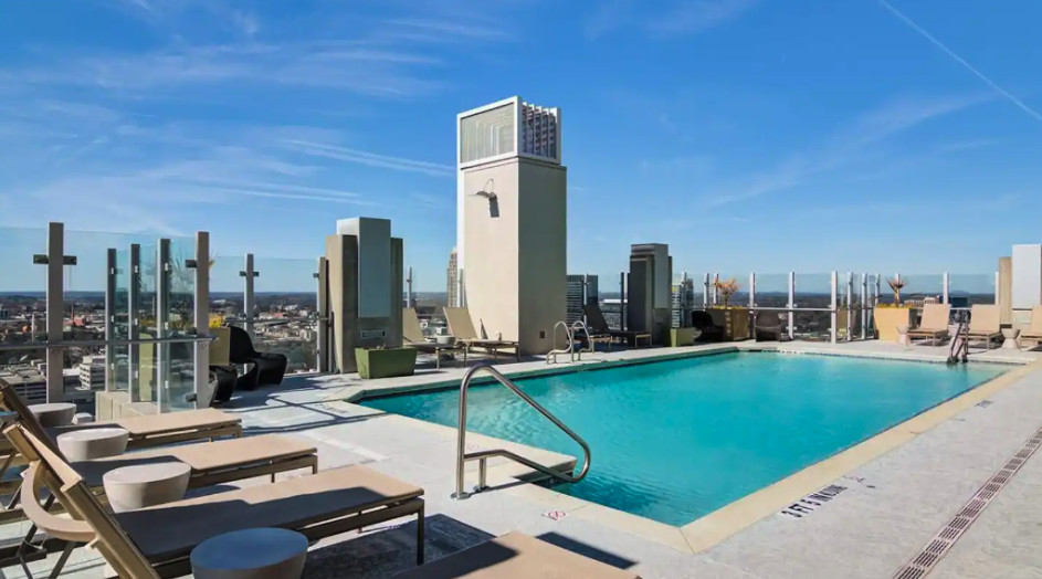 Rooftop swimming pool on a sunny day with city skyline views, lounge chairs, and glass railings.