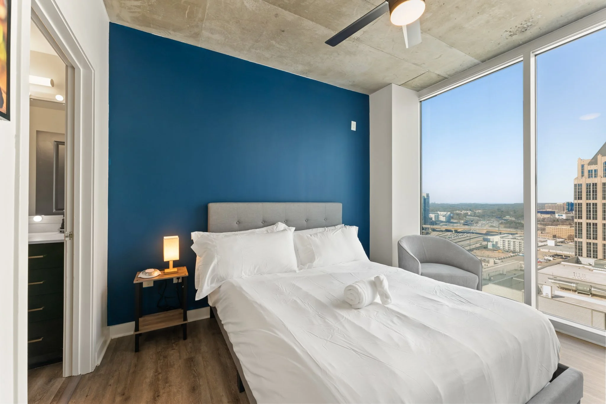 Modern bedroom with a large bed, white bedding, blue accent wall, small bedside lamp, window with city view, and a gray armchair.