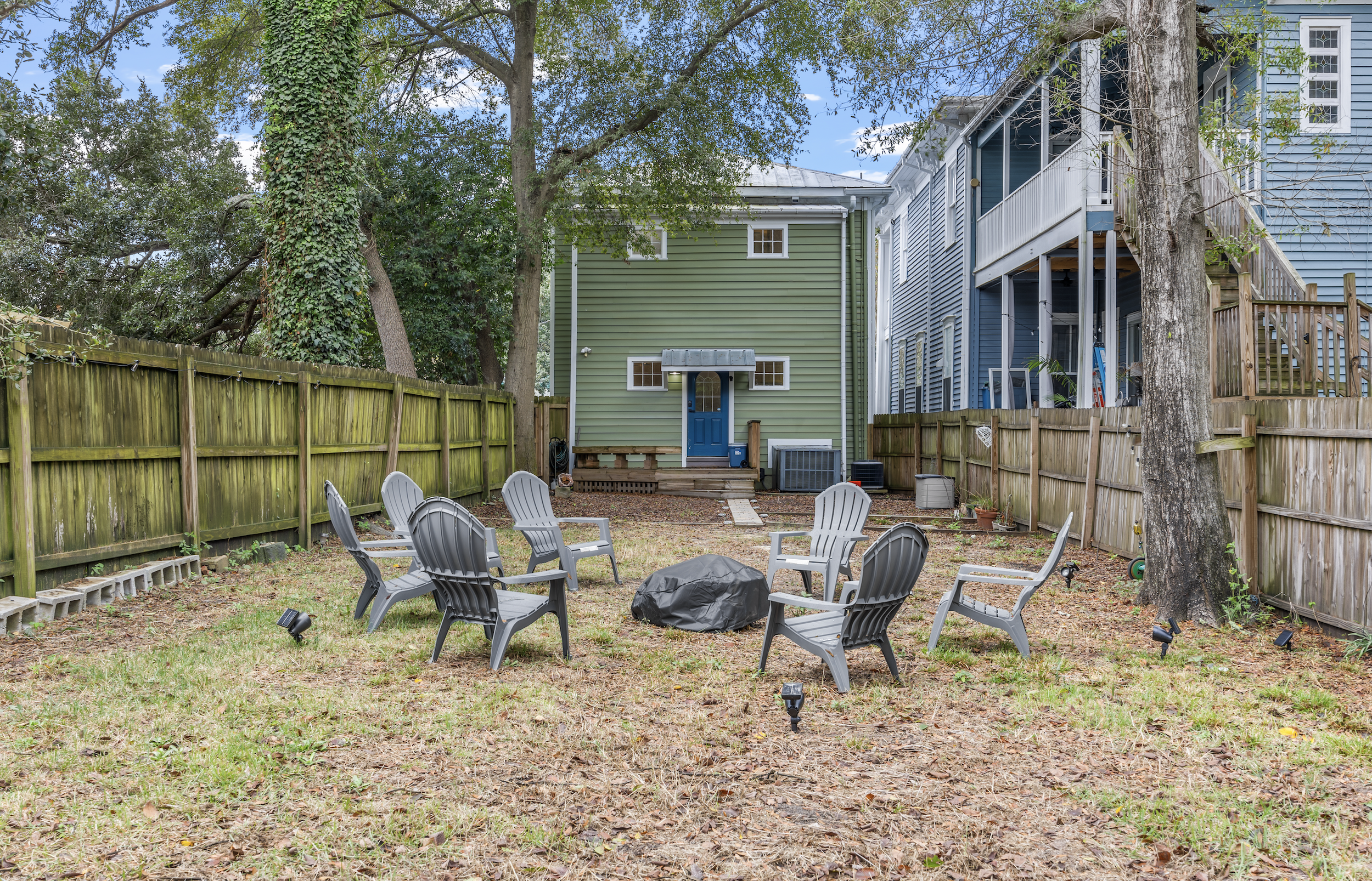 Backyard with six Adirondack chairs arranged in a circle around a covered object, possibly a grill, on a patchy lawn. A green house with a blue door and small porch is in the background, flanked by a tall tree on the right and a green wooden fence on