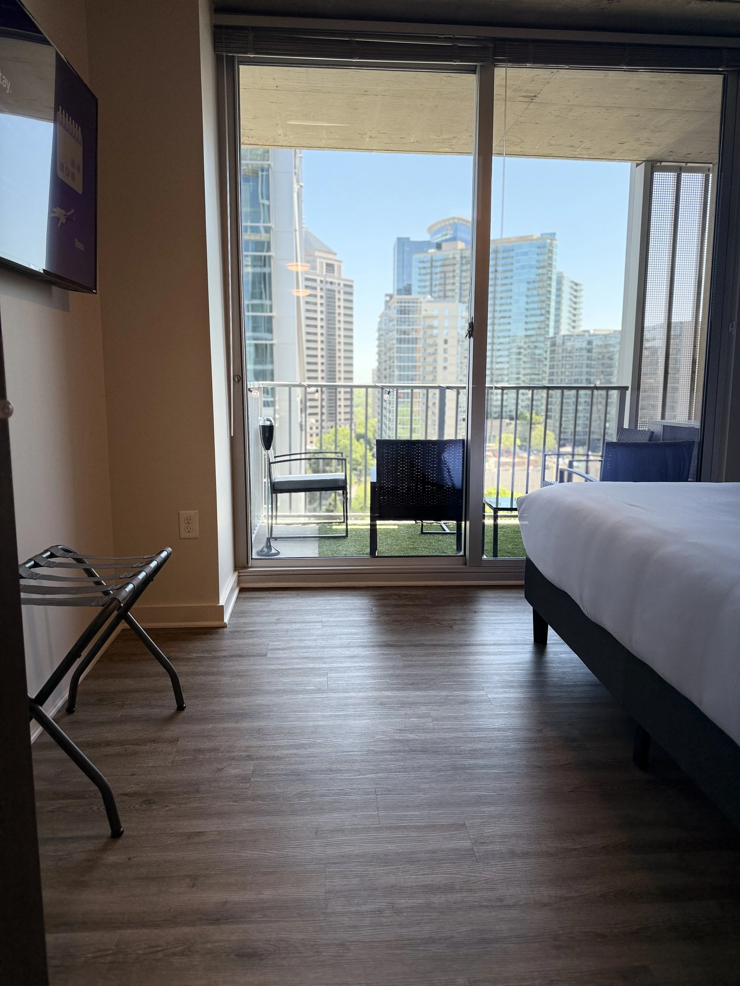 A hotel room with a sliding glass door leading to a balcony overlooking a city skyline of tall modern buildings. The room has wooden flooring, a bed with white linens, two chairs on the balcony, and a wall-mounted TV.