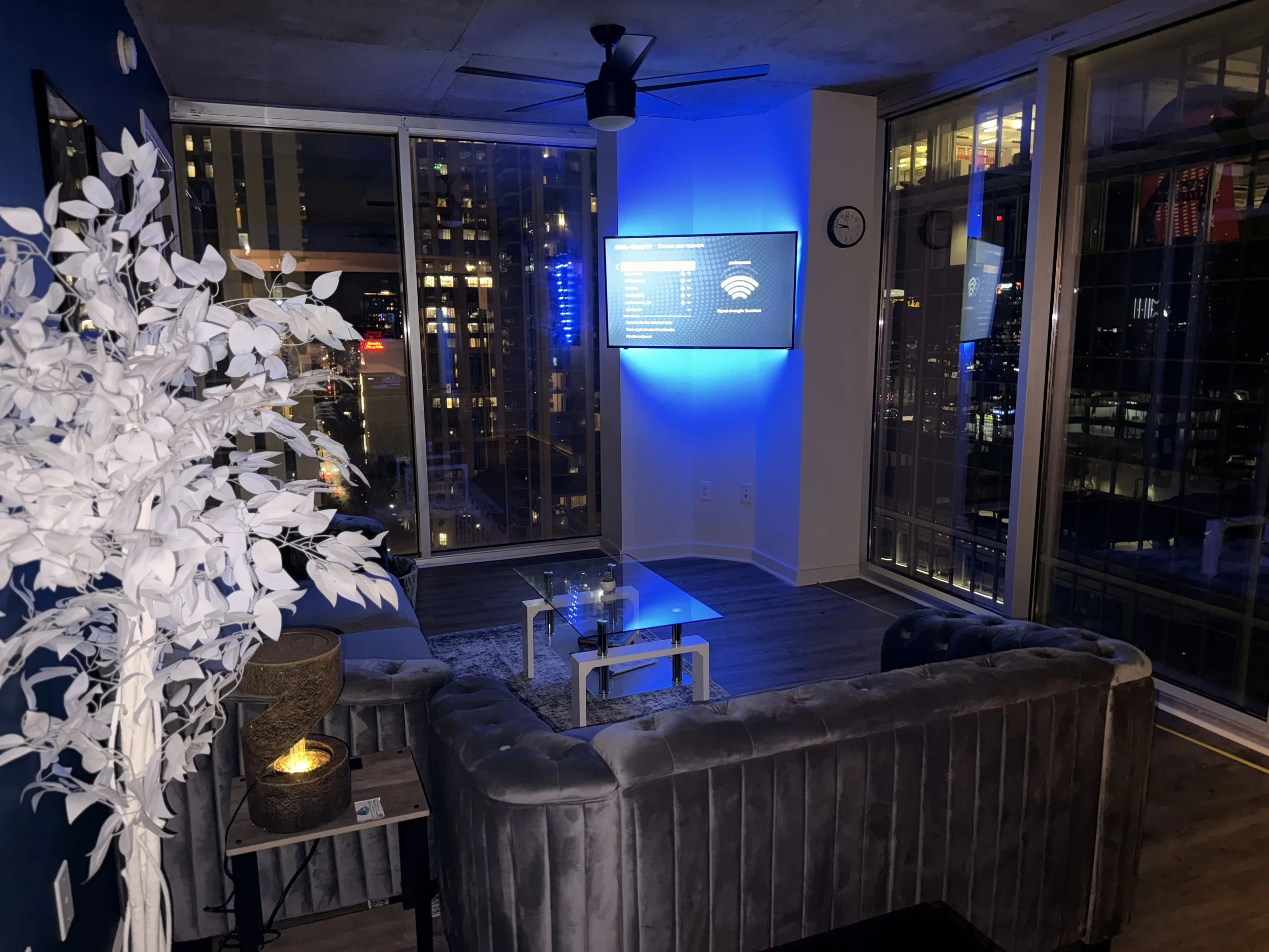 Modern apartment living room at night with large windows, city view, gray velvet sofa, glass coffee table, and a blue-lit TV wall.
