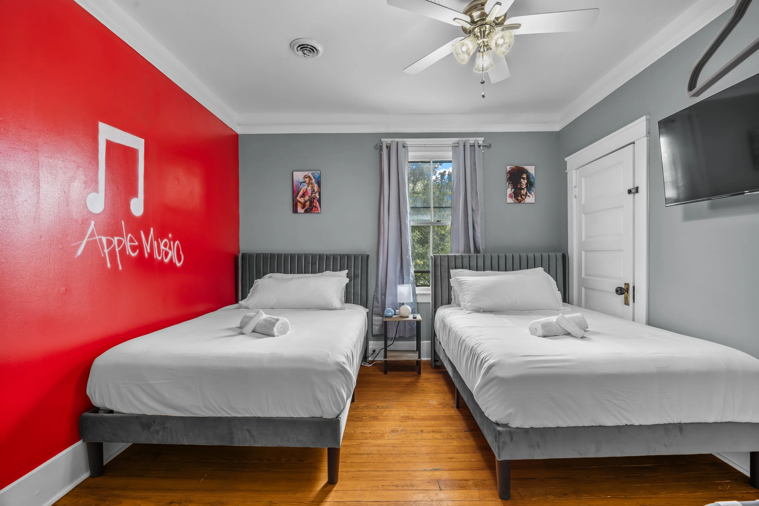 A bedroom with two twin beds with gray headboards, white bedding, and towels on each bed. One wall is painted red with the words 'Apple Music' and a music note logo. The other walls are light gray, with two colorful art posters, a window with curtain