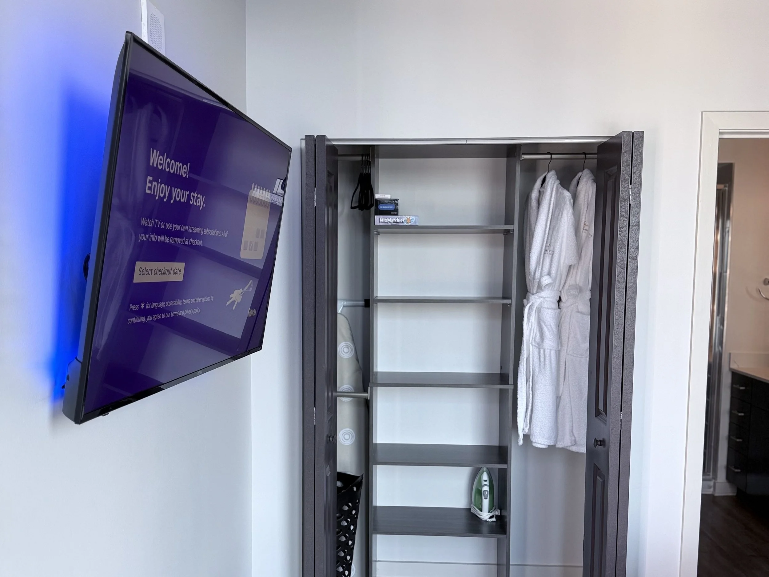Empty hotel closet with two white bathrobes hanging on the door, an iron on the bottom shelf, and a small device on the top shelf. A flat-screen TV mounted on the wall displays a welcome message.