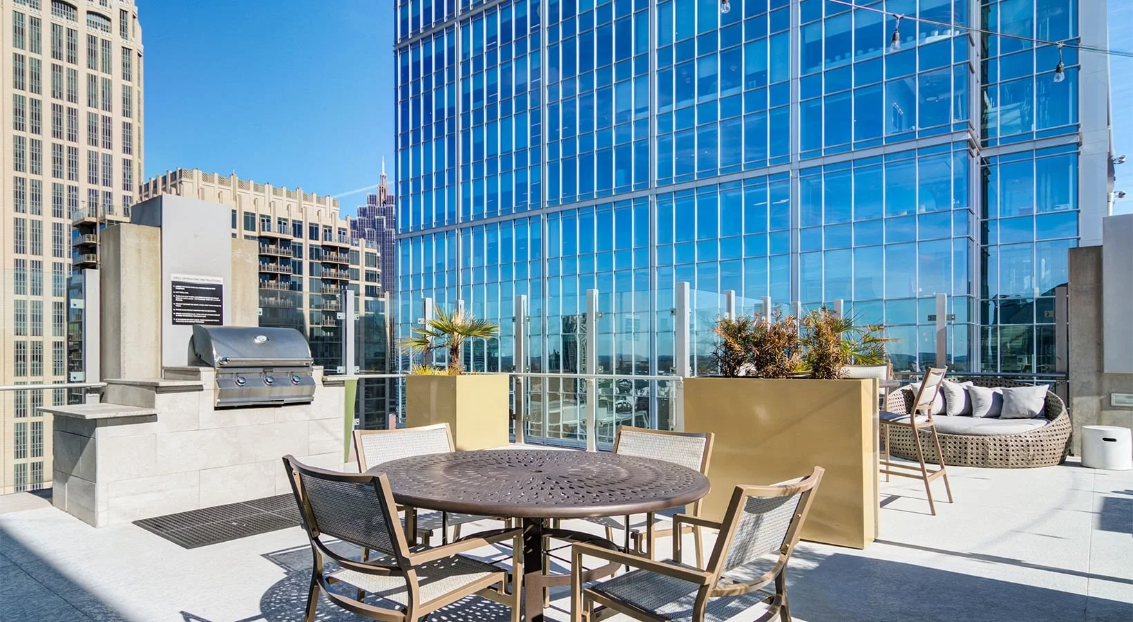 Rooftop patio with round table and chairs, outdoor sofa, potted plants, and city skyline with glass buildings in the background