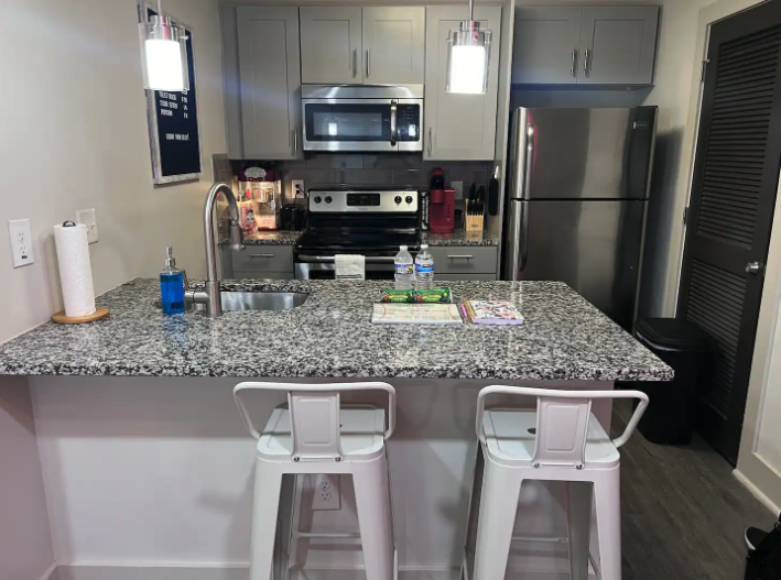 Modern kitchen with granite countertop, two white barstools, stainless steel refrigerator, microwave, and stove, with bottles of water and kitchen utensils on the counter.