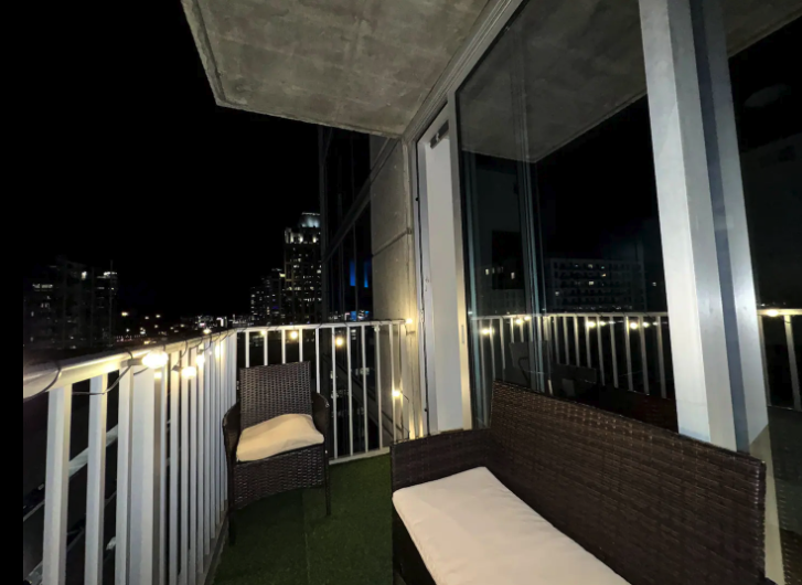 A balcony at night with two woven outdoor chairs with cushions, string lights along the railing, and city buildings with illuminated windows in the background.