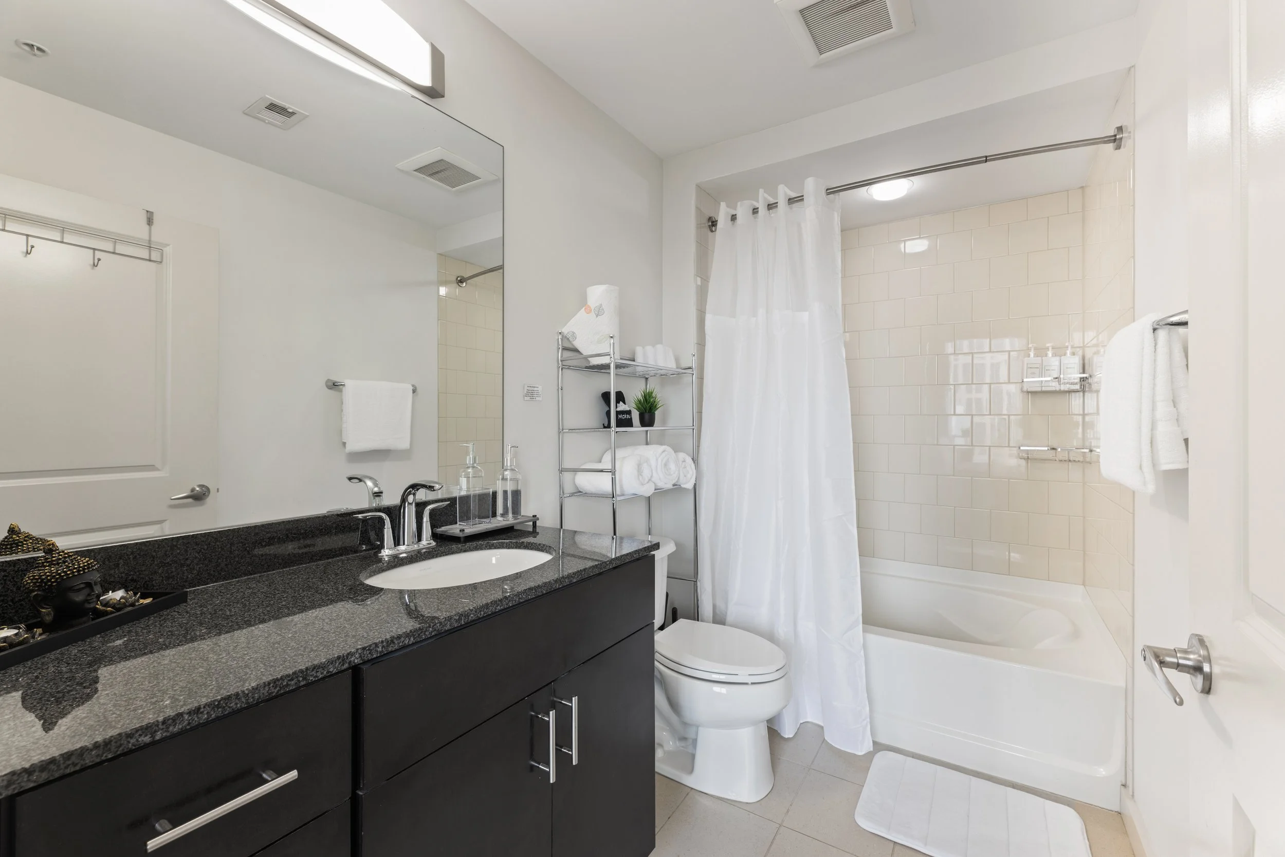Modern bathroom with dark vanity, large mirror, white shower curtain, bathtub, white tiles, and bathroom accessories.