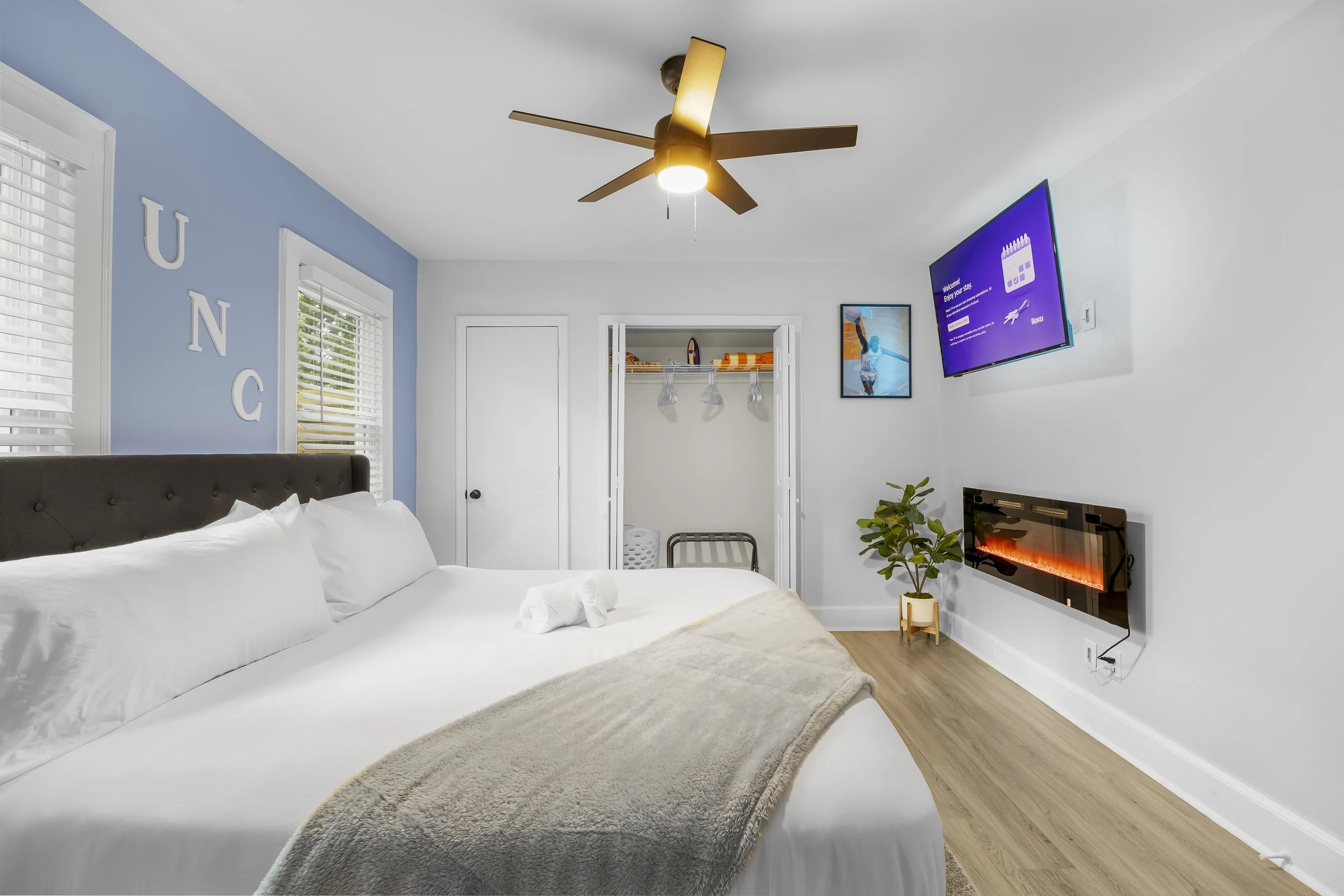 Modern bedroom with a large bed, white bedding, and a beige throw blanket. Blue accent wall with the word 'UNC' in white letters. Two windows with white blinds, a ceiling fan, and a flat-screen TV mounted on the wall. A decorative electric fireplace 