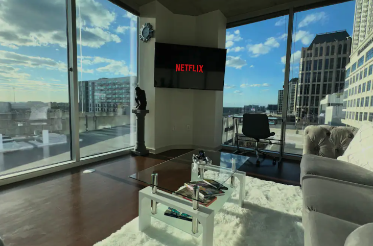 Modern city apartment living room with large windows, a wall-mounted Netflix screen, a glass coffee table with books, a white rug, and a mix of contemporary furniture, overlooking a city skyline.