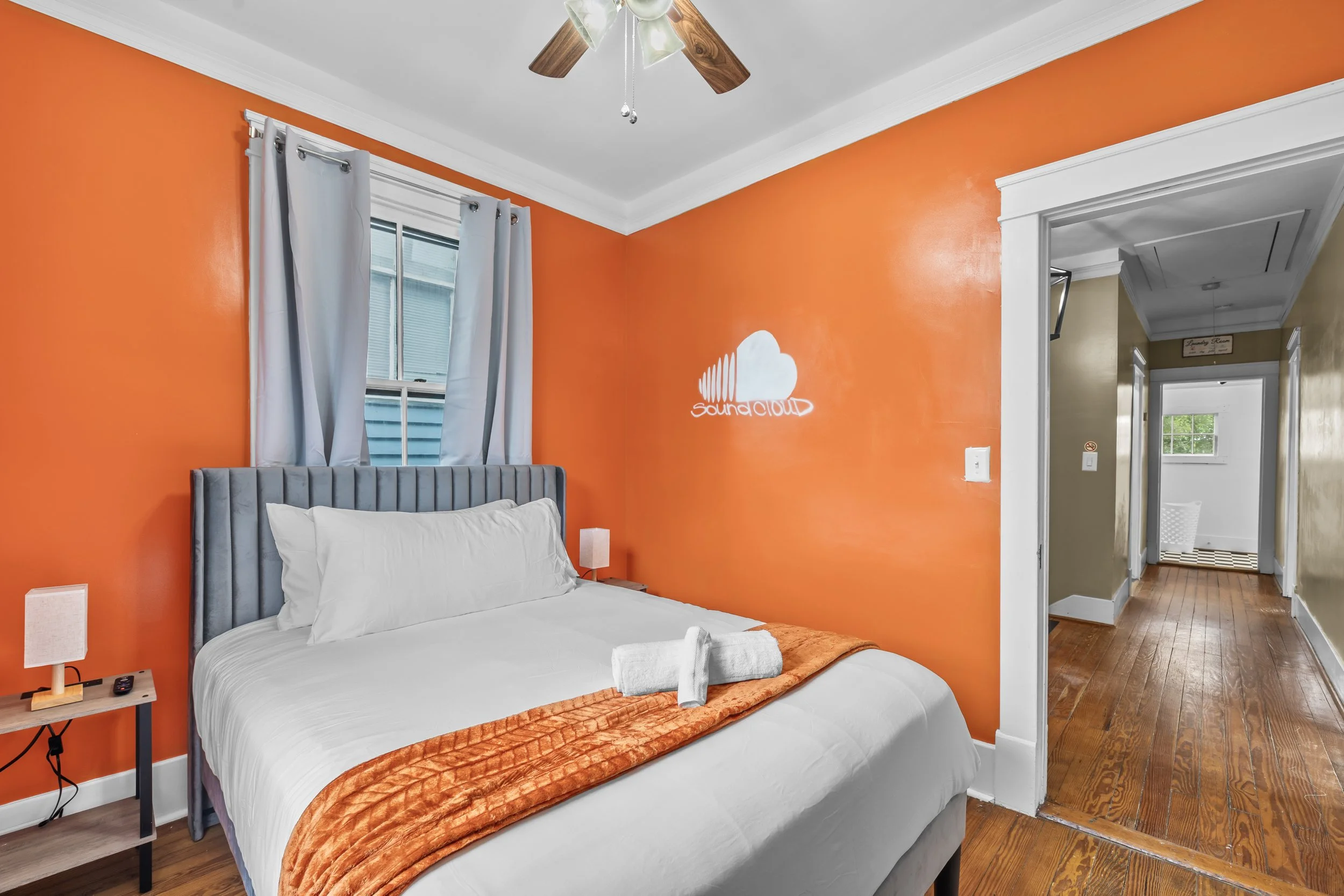 Bedroom with orange accent wall, gray upholstered bed with white bedding, small nightstands with lamps, window with white curtains, hallway leading to other rooms, wooden floors, sound cloud mural on wall.
