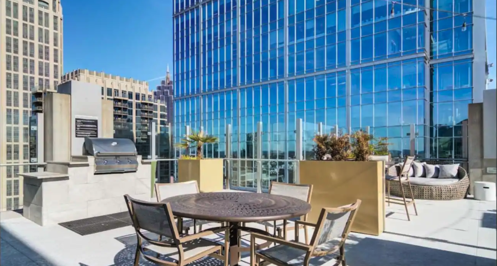 Rooftop terrace with outdoor dining table, chairs, a grill, and lounge seating, with modern blue glass buildings in the background on a clear day.