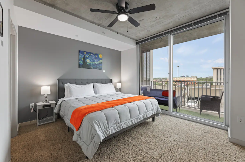 Modern bedroom with a king-sized bed, gray headboard, white bedding, orange throw blanket, two bedside lamps, artwork on gray wall, ceiling fan, and sliding glass door leading to a balcony with outdoor seating and city view.
