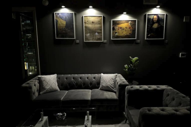 A dimly lit living room with a gray tufted sofa, matching armchair, and a glass coffee table. Wall art includes four framed paintings, and a potted plant is nearby. There is a refrigerator or display case on the left.