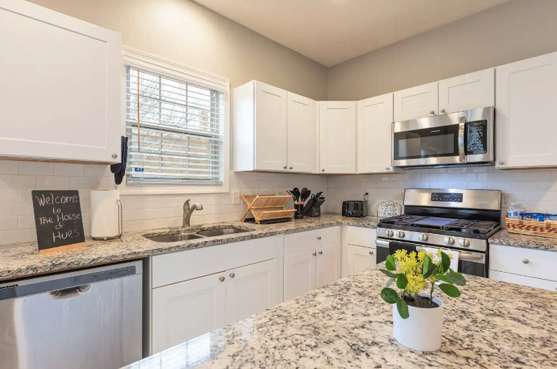 Kitchen with white cabinets, granite countertops, stainless steel microwave and stove, window above the sink, small chalkboard sign, and a potted plant on the island.