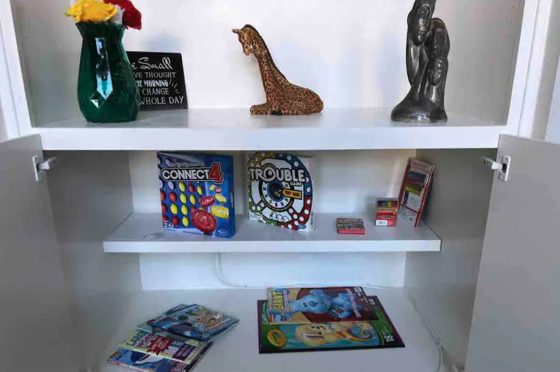 White cabinet with two shelves containing board games, books, and small toys. Decor includes a green vase with flowers, a giraffe figurine, and a sculpture of a woman. The top shelf also has a black sign.