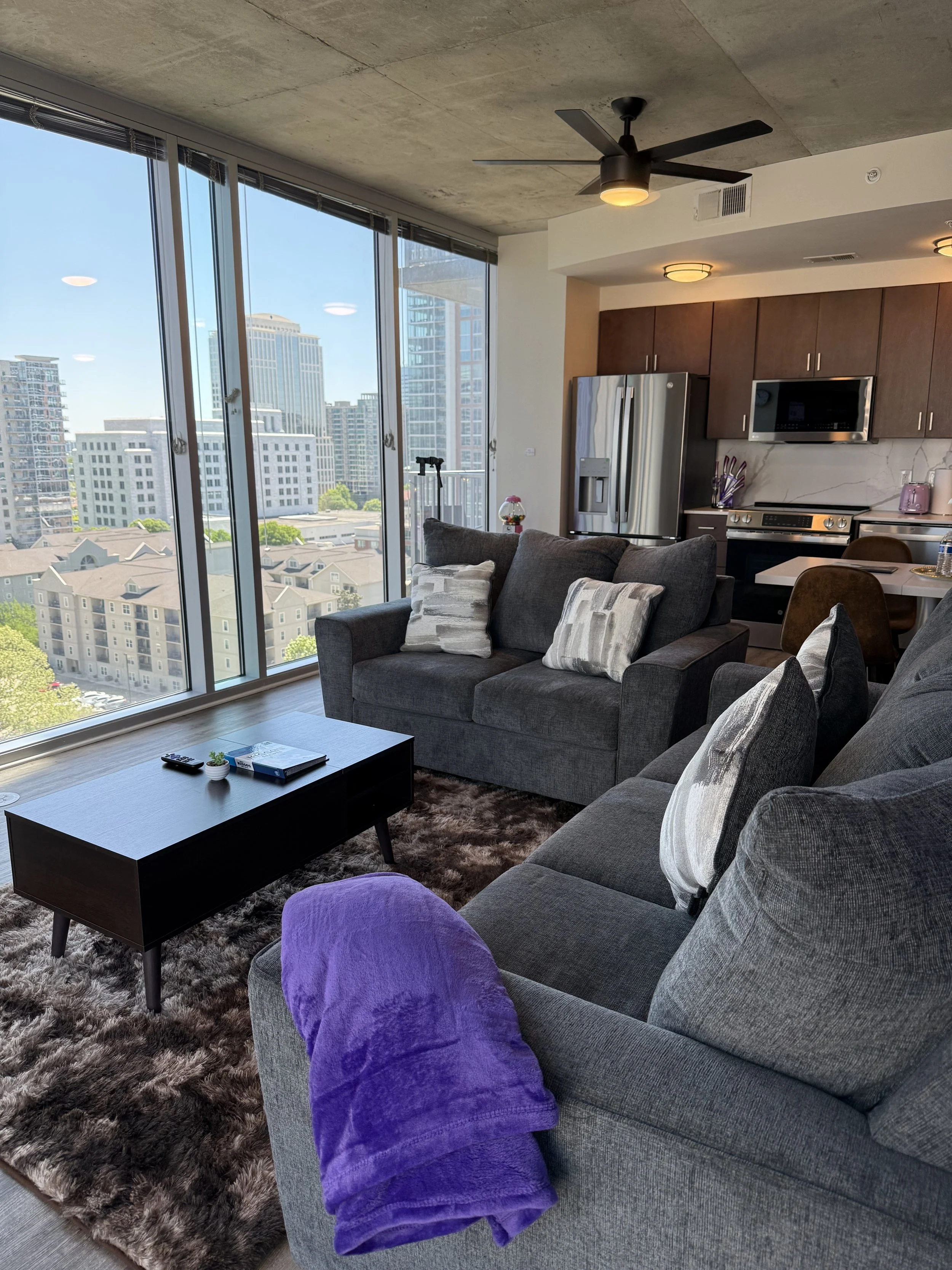 Living room with large floor-to-ceiling windows showing city skyscrapers, two gray sofas with pillows, brown shag rug, black coffee table with remote and small potted plant, purple blanket on one sofa, kitchen in background with stainless steel fridg