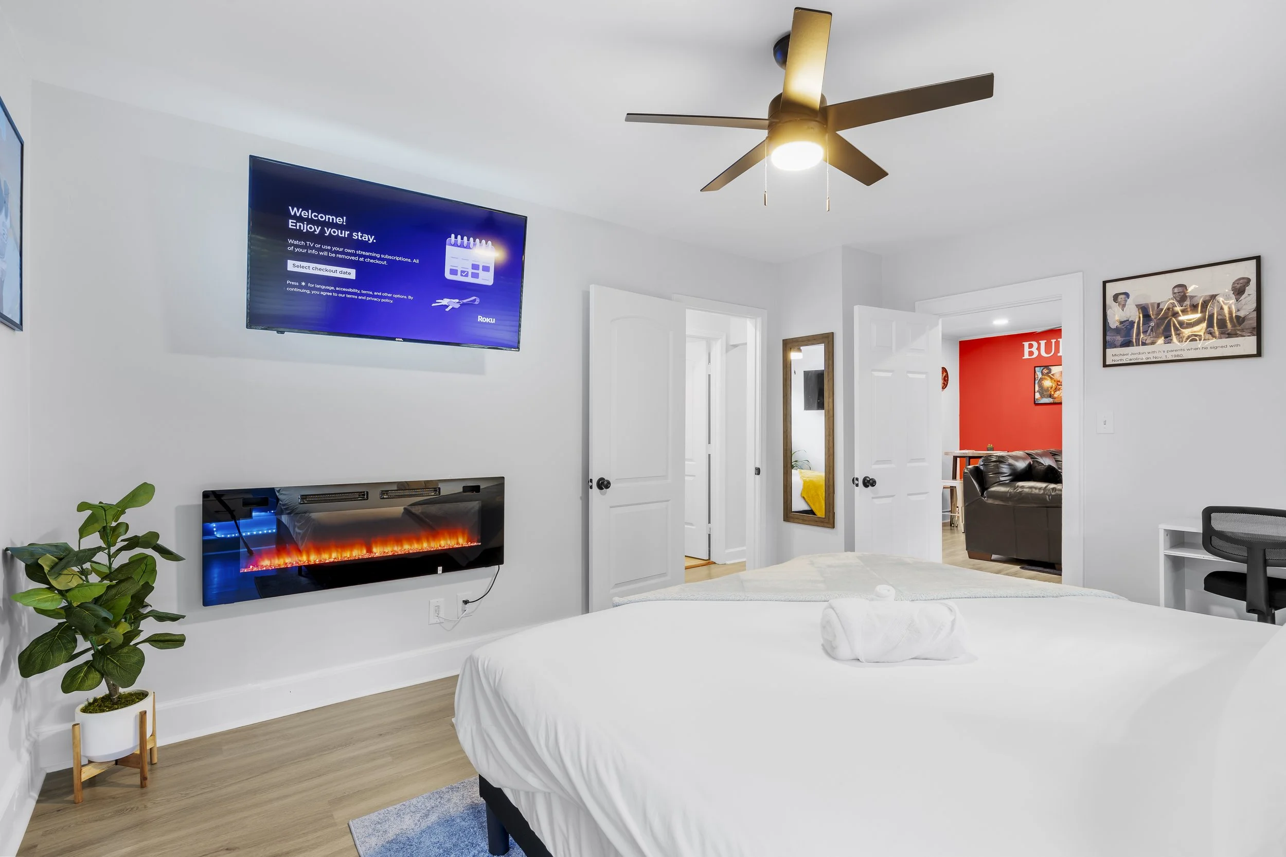 A bedroom with a white bed, a potted plant, a wall-mounted television, a modern electric fireplace, and a ceiling fan. An open door reveals part of another room with red walls and a black couch.