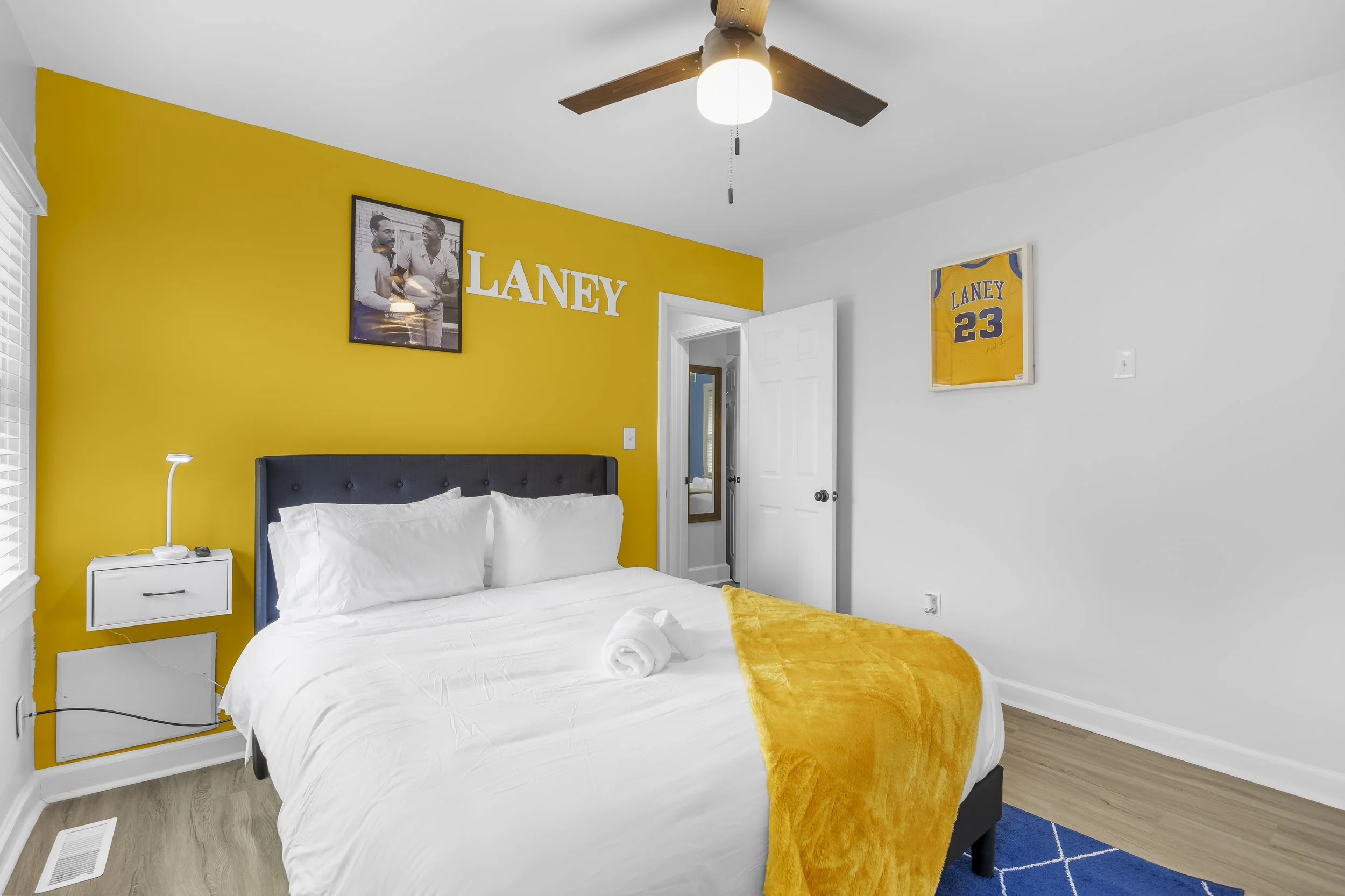 Bedroom with a yellow accent wall, framed sports memorabilia, a bed with white linens and a yellow throw, and a ceiling fan with wooden blades.