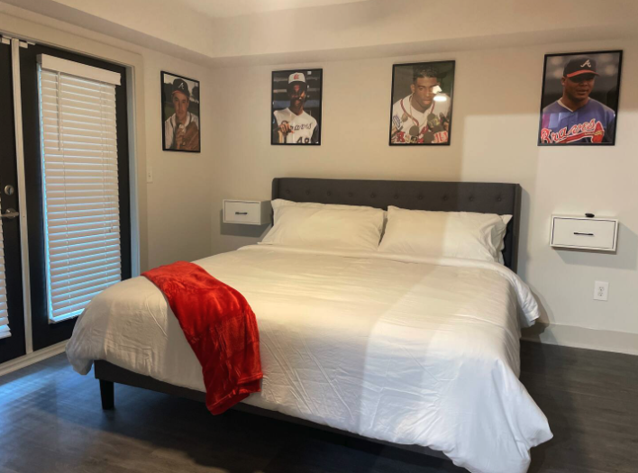 A bedroom with a large bed dressed in white bedding, a red blanket at the foot of the bed, and three framed sports photos above the headboard. There are two small wall-mounted nightstands and a sliding glass door with blinds.