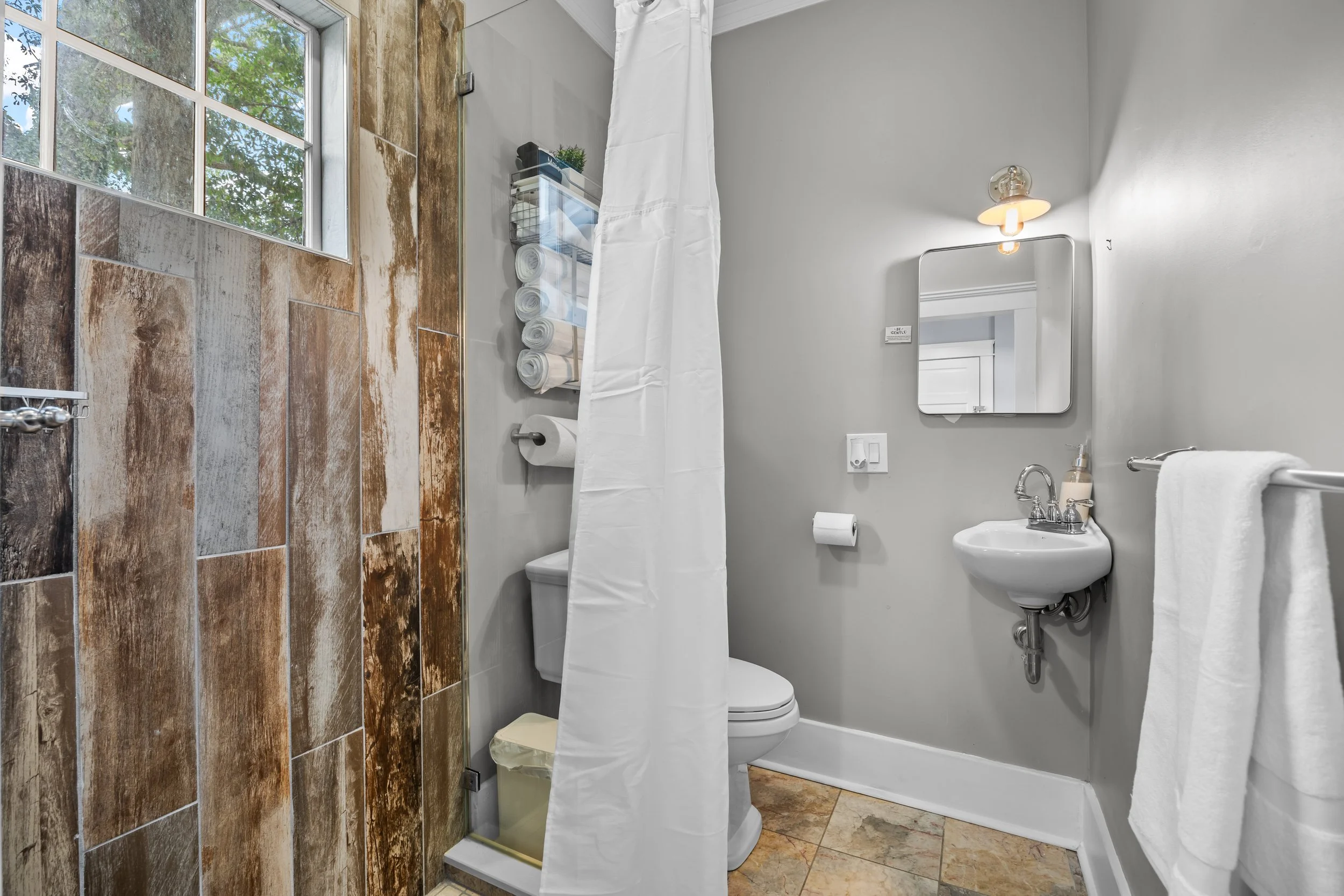 Small bathroom with a window, tile floor, gray walls, a white toilet, a small wall-mounted sink, a mirror, a towel rack with a white towel, and a shower area with a brown-toned tile wall, a curtain, and a shelf with rolled towels.