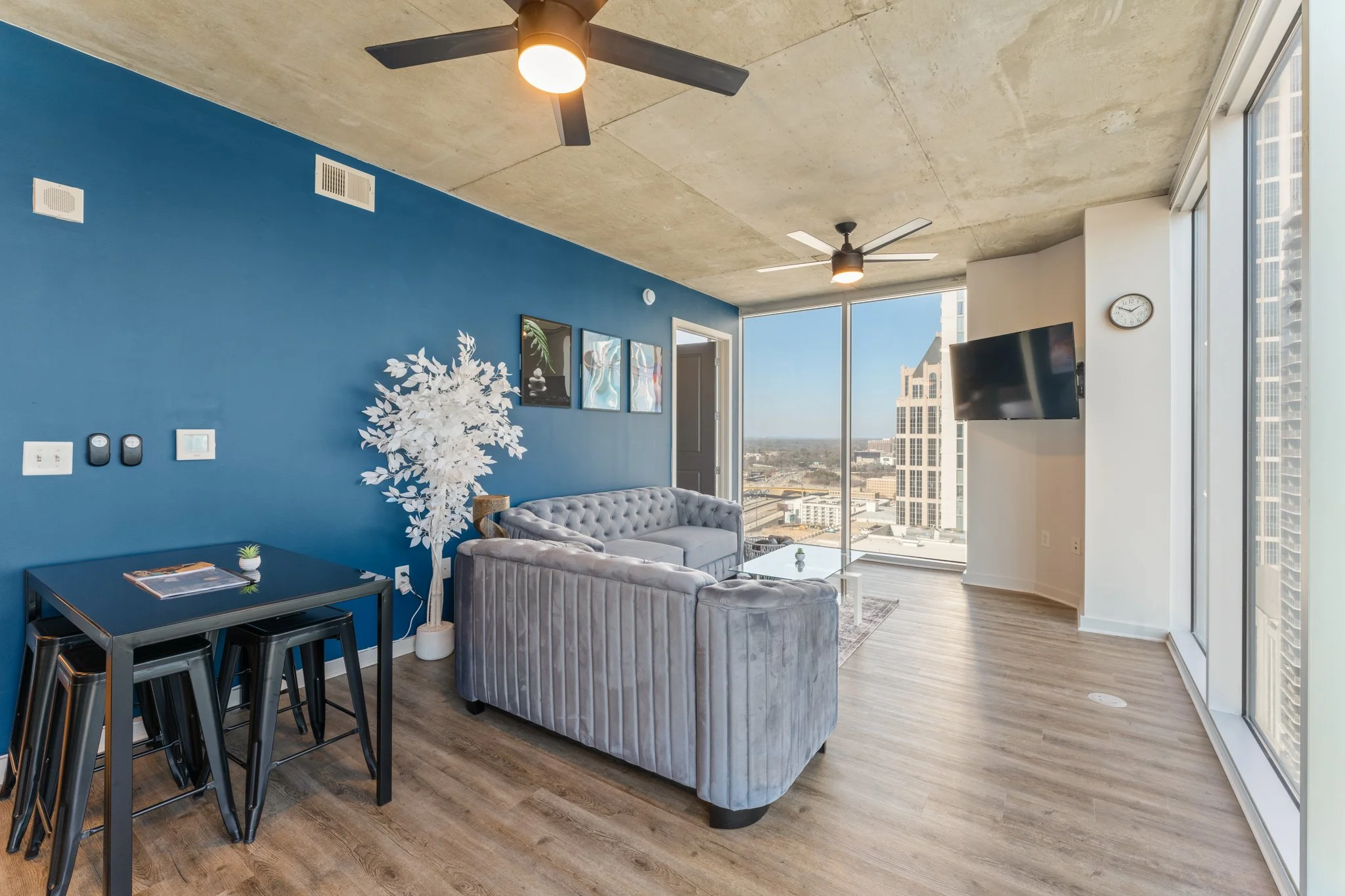 Modern living room with large window view, blue accent wall, gray velvet sofa, black table with stools, white decorative tree, wall art, and ceiling fans.