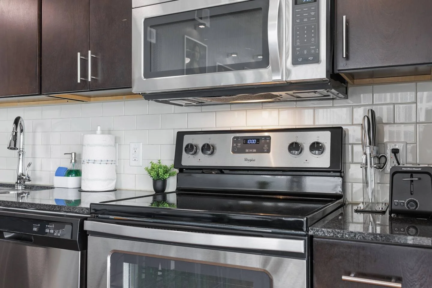 Modern kitchen with stainless steel microwave over oven, black and stainless steel stove, white subway tile backsplash, dark wood cabinets, granite countertop, and various small appliances including a toaster, and cleaning supplies on the counter.