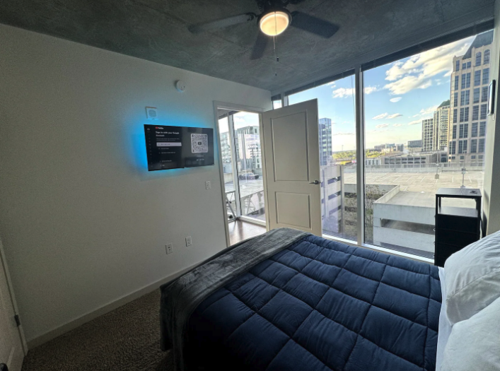 Modern bedroom with large windows showing city view, bed with navy blue quilt, wall-mounted TV, and ceiling fan.
