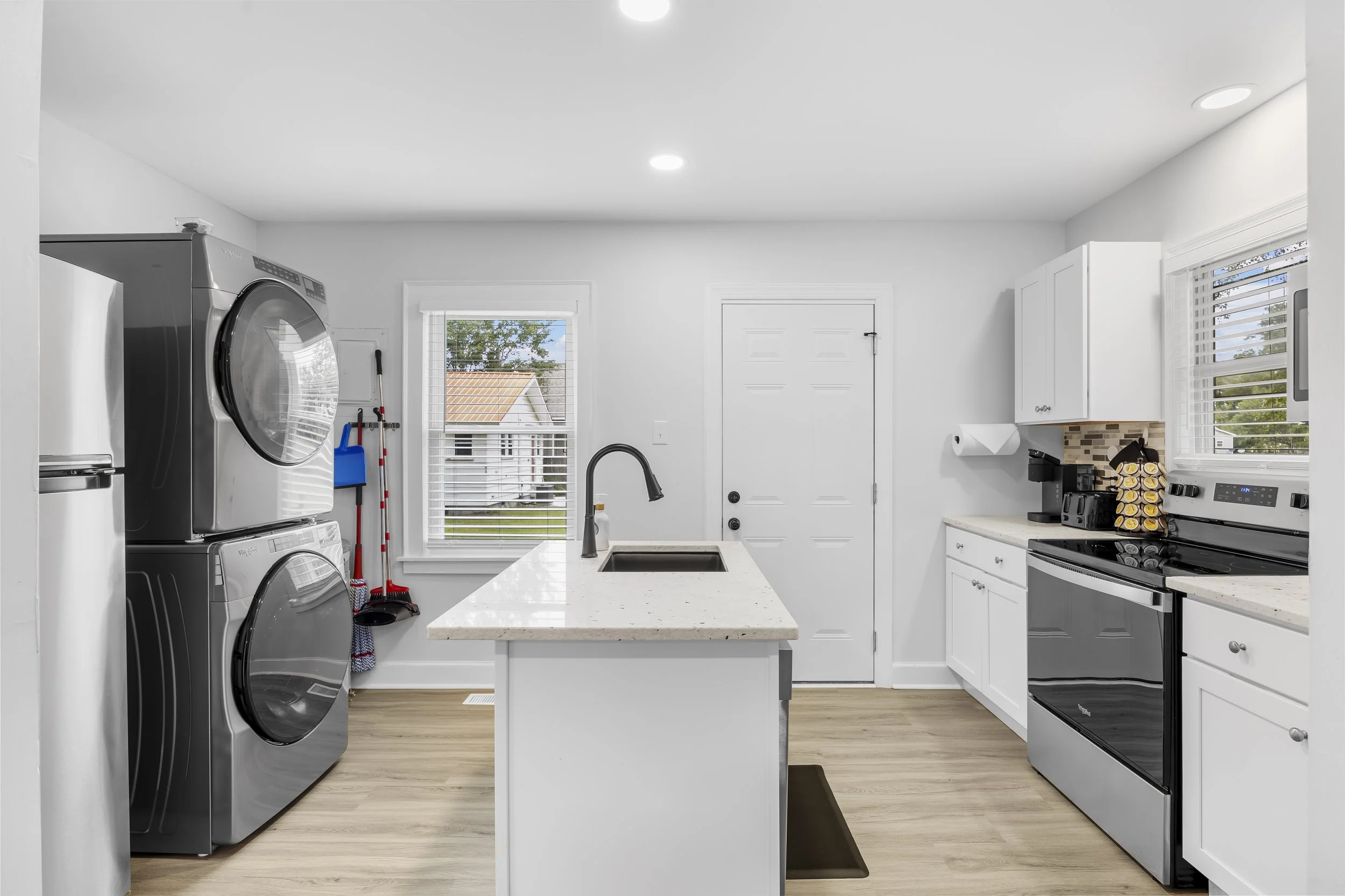 Modern laundry room with stacked front-loading washer and dryer, white kitchen cabinets, a black stove, a small kitchen island with a sink, and a window showing a neighborhood outside.