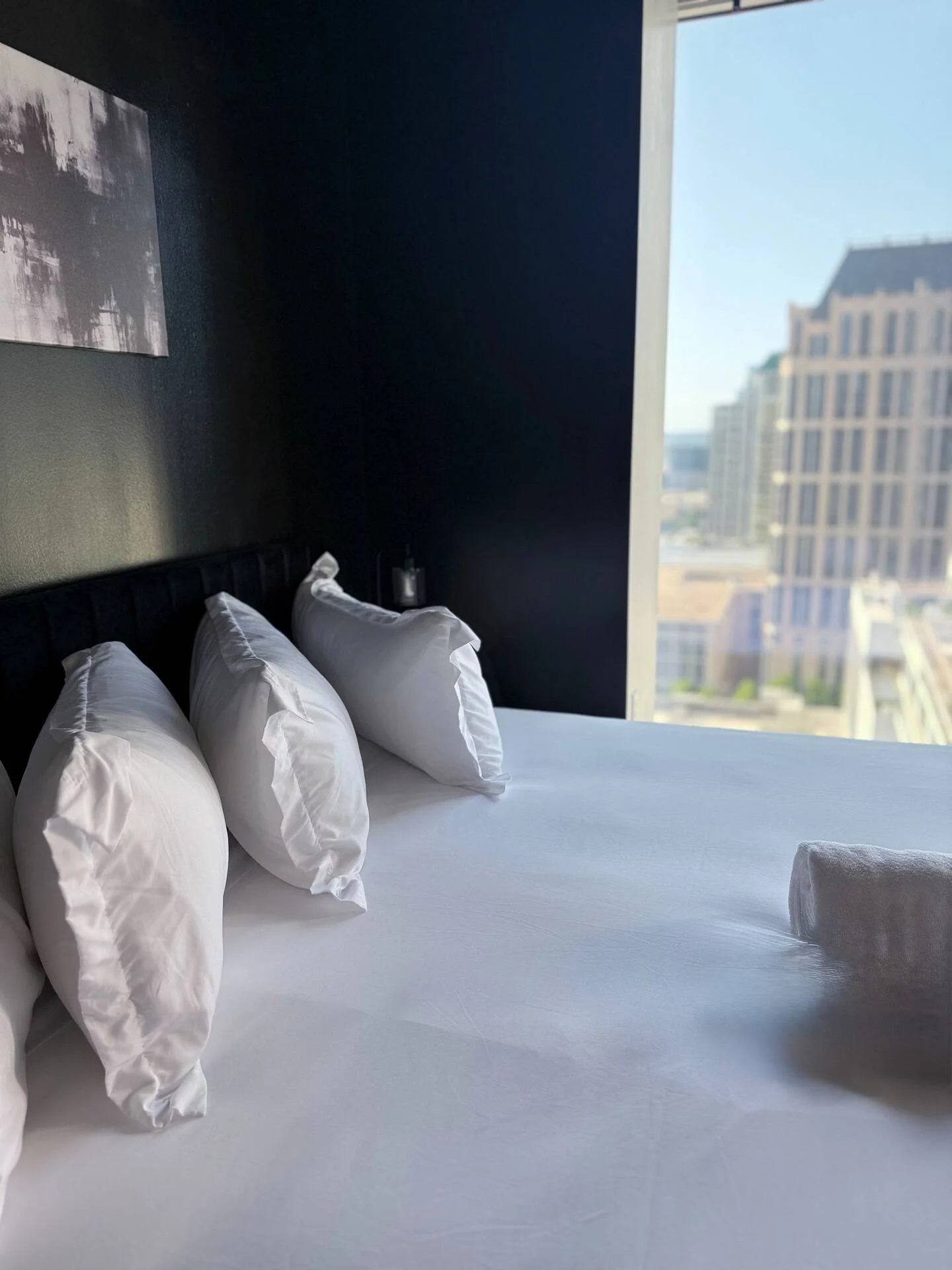 A neatly made bed with four piled white pillows and a rolled towel at the end, in a modern hotel room with dark walls and a large window showing a cityscape.