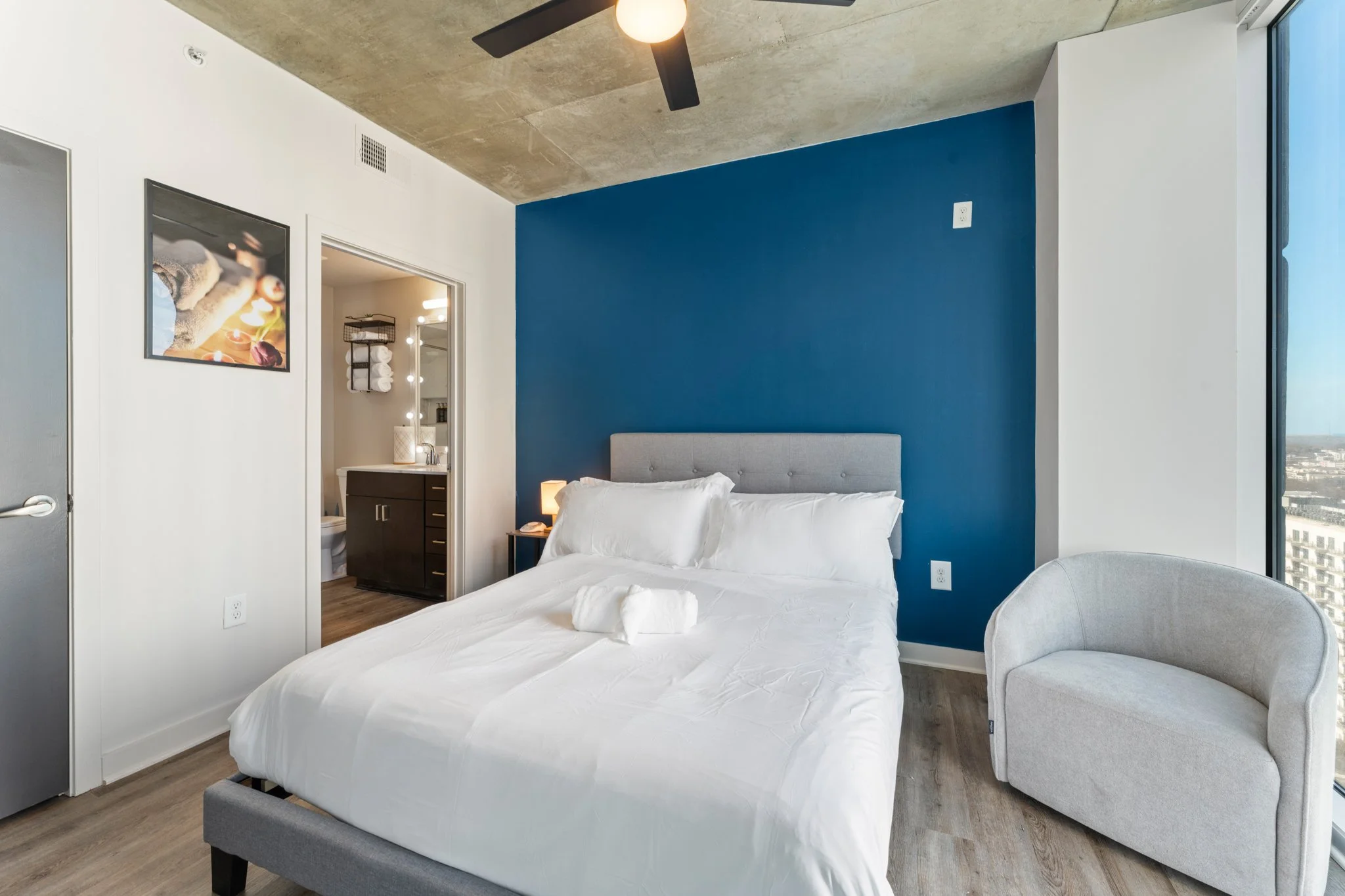 Modern hotel room with a white bed, gray headboard, blue accent wall, beige armchair, bedside lamp, and a large window showing cityscape.