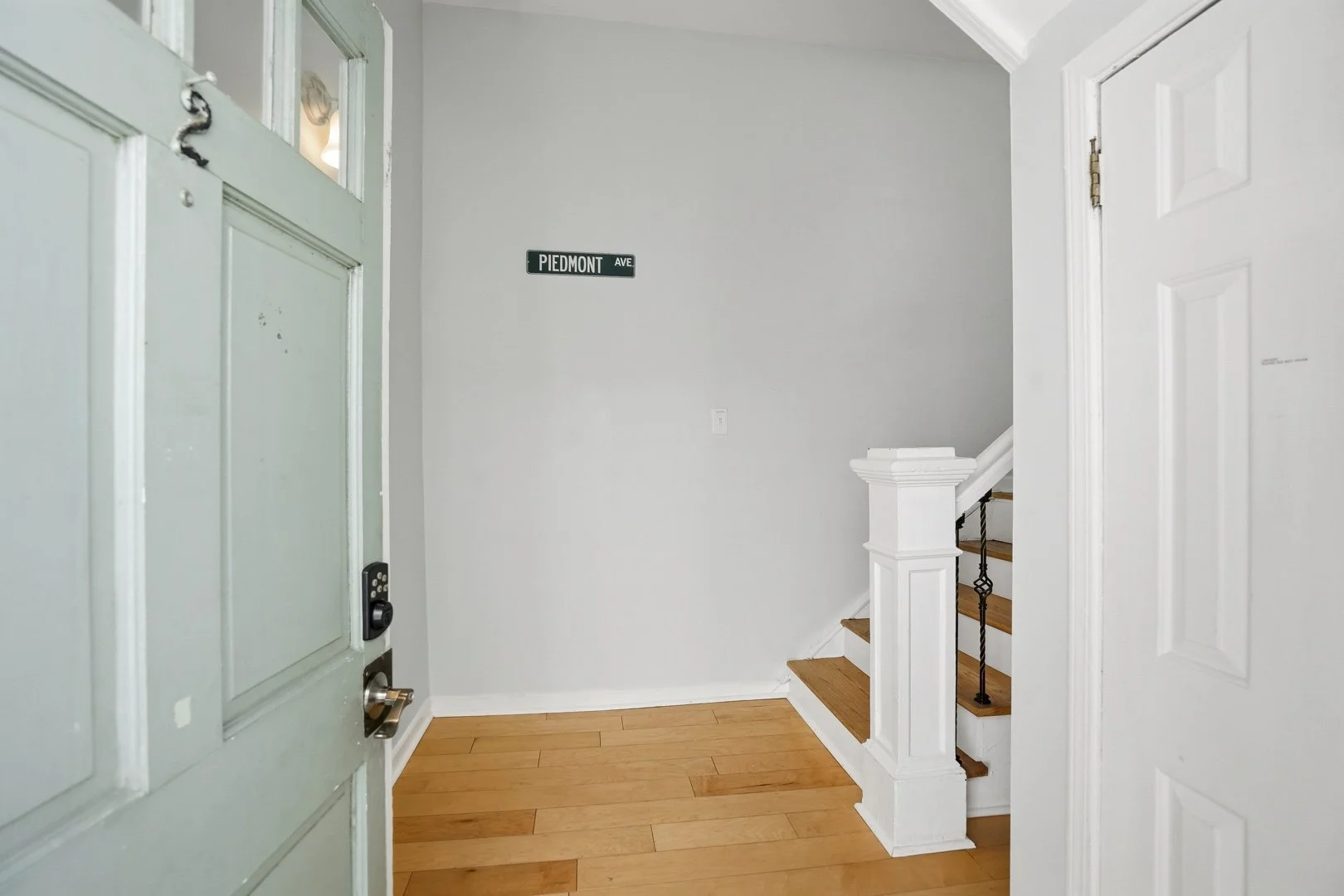 Entryway with hardwood floor, white walls, staircase with black iron railing, and a small street sign reading "Piedmont Ave" on the wall.