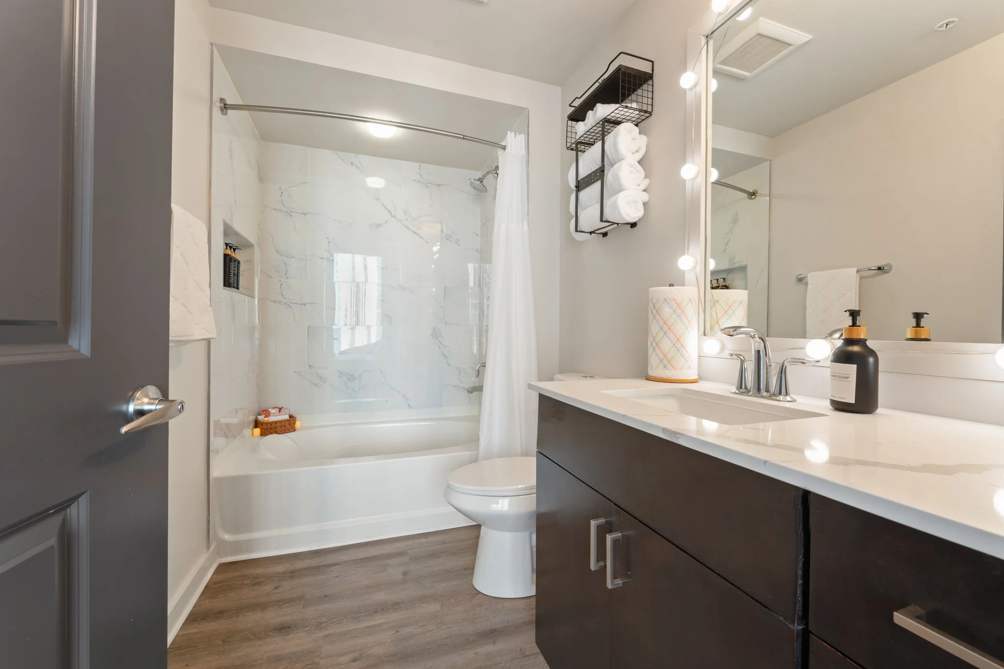 A clean, modern bathroom with a bathtub and shower, a toilet, a dark wood vanity with a white marble countertop, and a large mirror framed with lights.