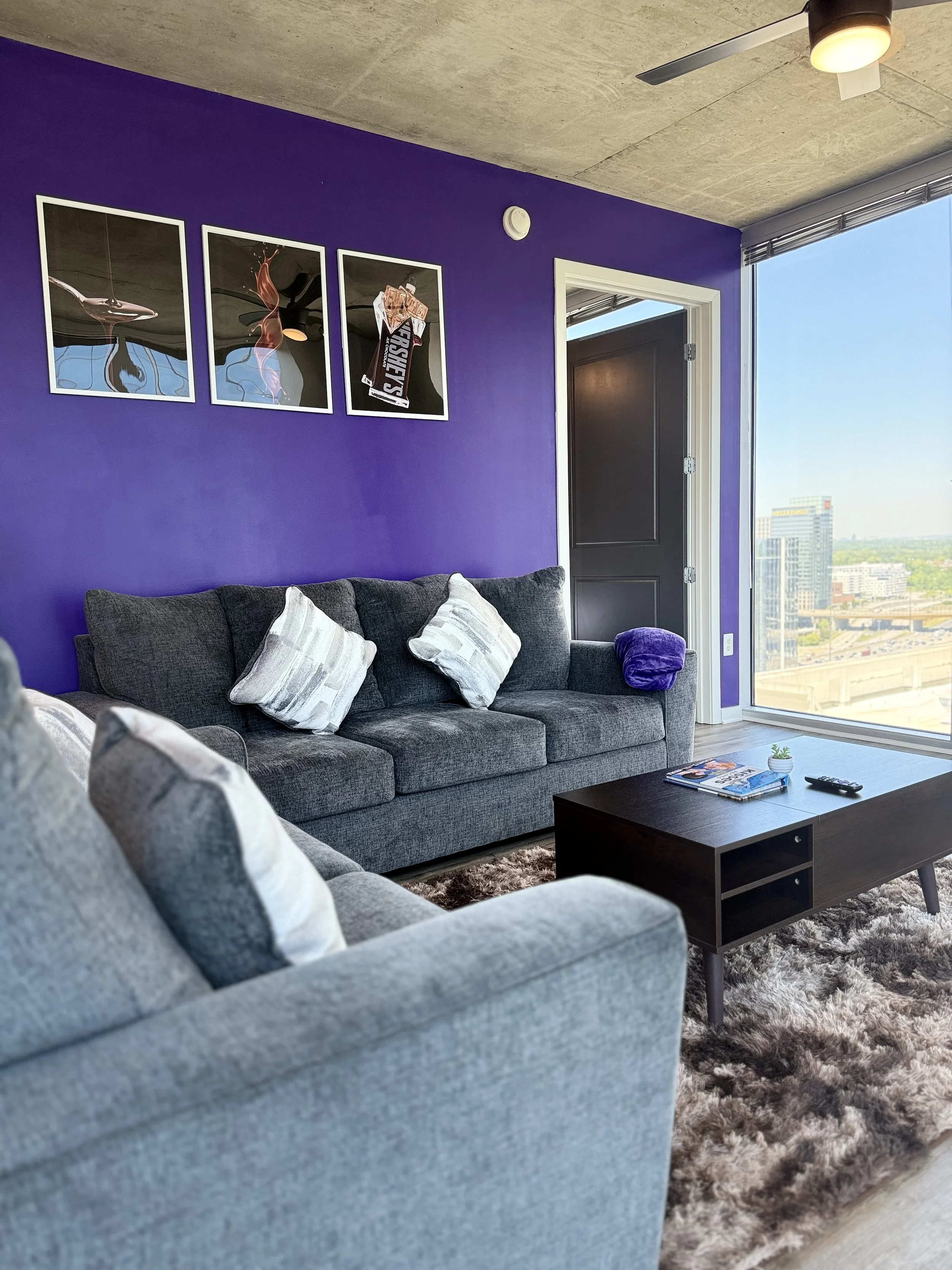 Modern living room with purple accent wall, gray sofa with white pillows, dark wooden coffee table, and large window revealing cityscape view.
