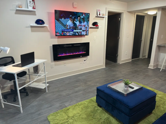 Living room with a mounted sports game on a TV, electric fireplace below, white desk with a laptop, blue ottoman on a green rug, and decorative shelves with hats and small decor items.