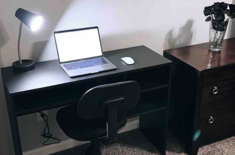 A black desk with a laptop, a white mouse, and a desk lamp, positioned next to a wooden dresser with a glass vase of black flowers on top.