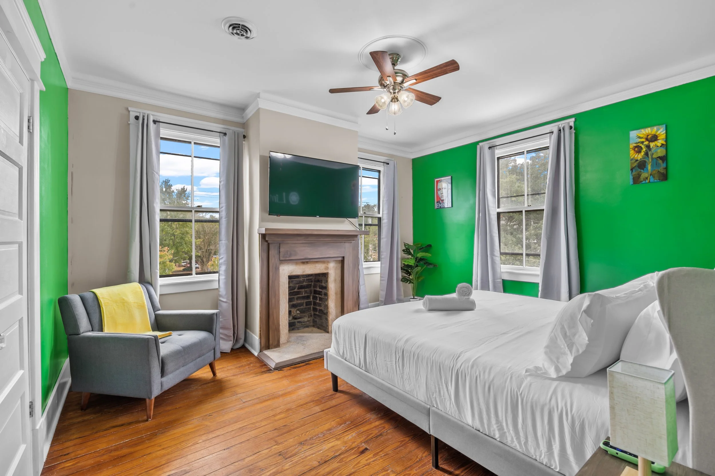 Bedroom with a white bed, gray armchair with yellow throw, green accent wall, white curtains, wooden floor, ceiling fan, fireplace with TV above, and windows showing trees outside.