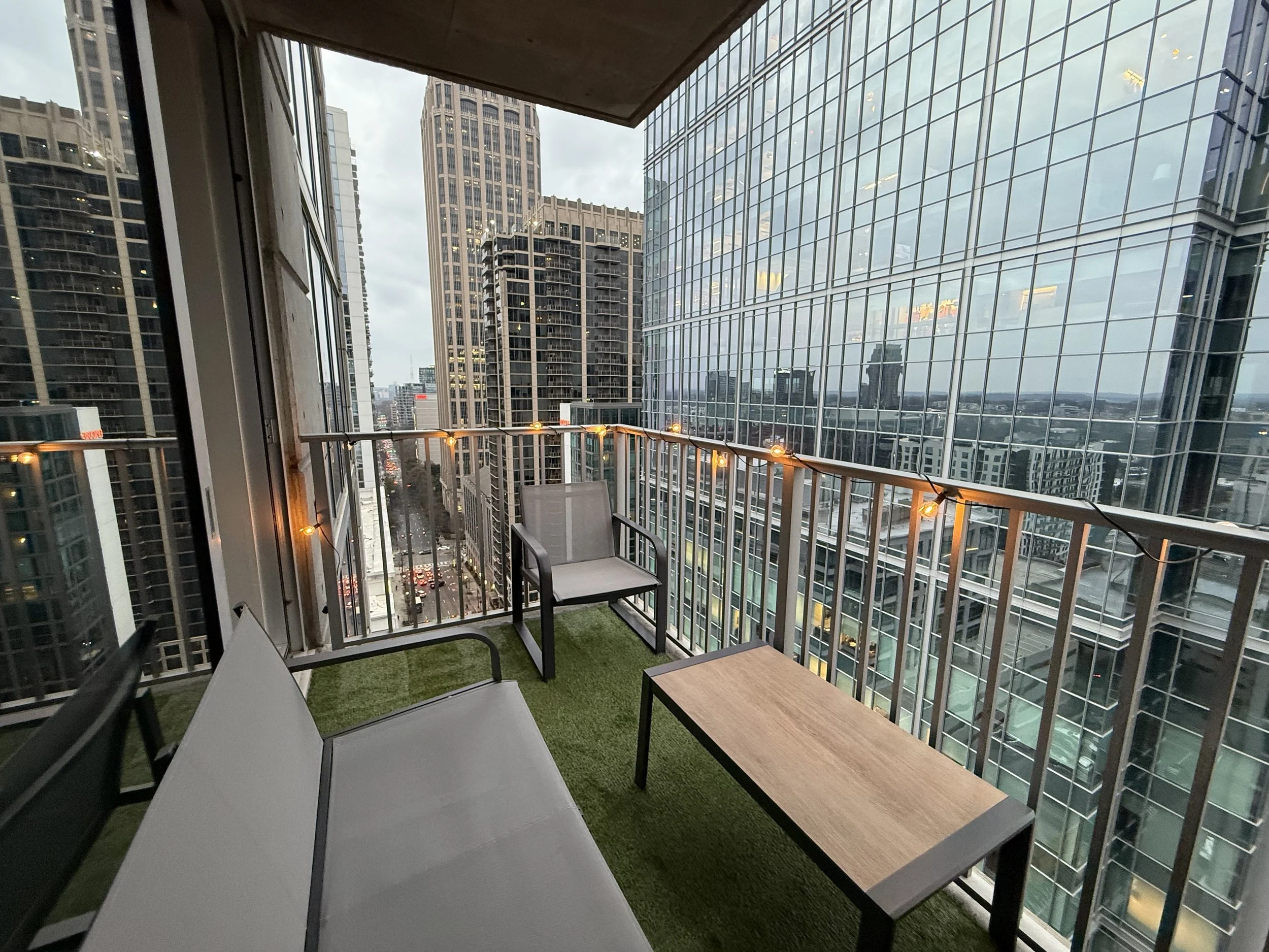 Balcony with string lights, chairs, and a small table overlooking a city with tall glass buildings
