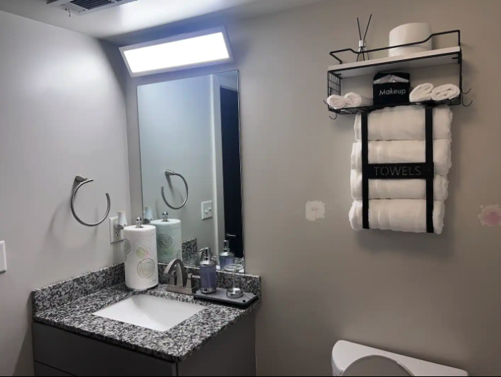 Bathroom with granite countertop sink, mirror, towel rings, and wall-mounted rack holding rolled towels, a makeup box, and a container. The wall has a light difference near the middle and pink spots.
