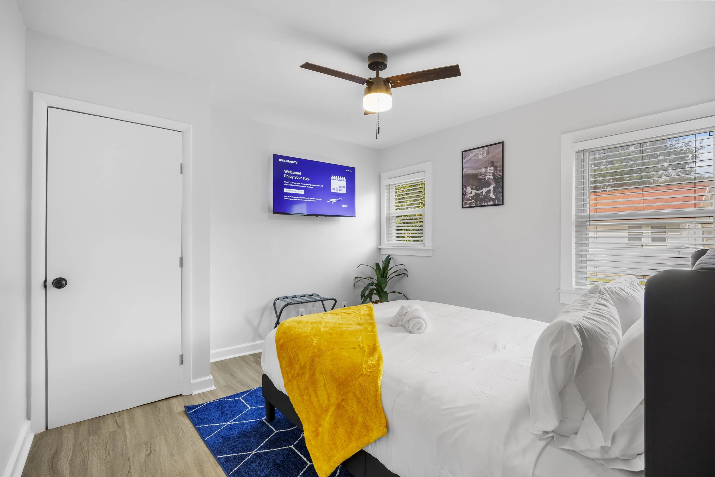 A bedroom with white walls and light wood flooring, featuring a bed with white bedding and a yellow blanket. There is a window with white blinds on the right, a wall-mounted TV on the left, a ceiling fan with wooden blades, and a small potted plant n