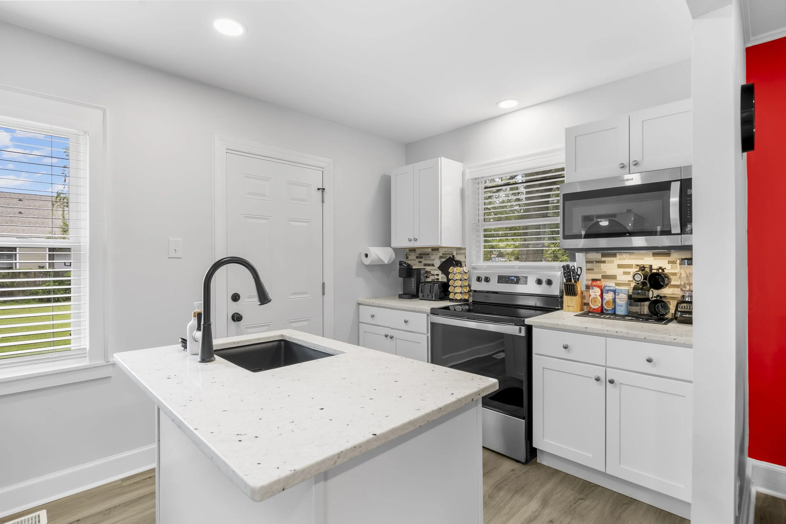 Modern kitchen with white cabinets, stainless steel stove, microwave and sink, island with white speckled countertop, red accent wall, window with blinds, and various small appliances and items on the counter.