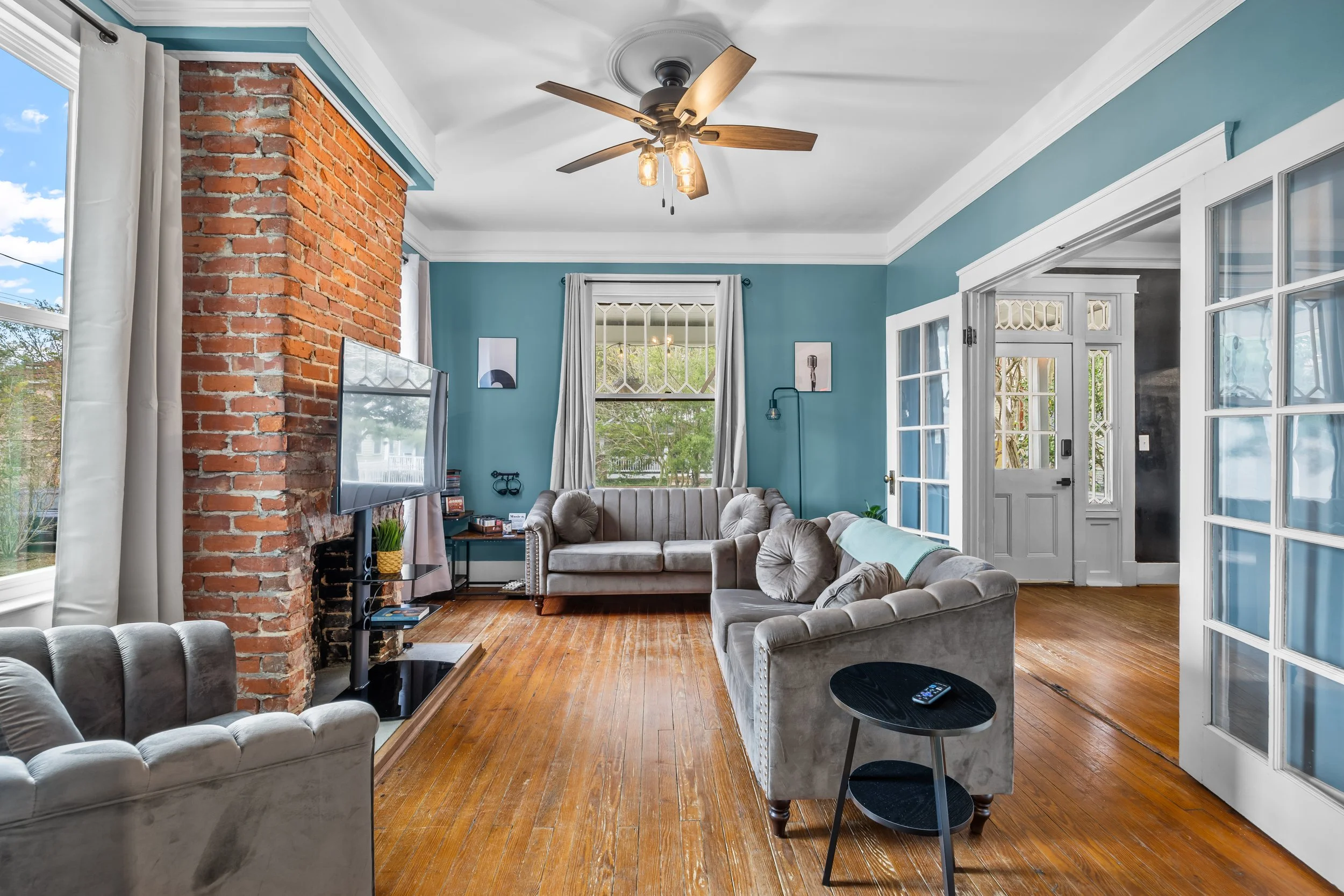Living room with blue walls, brick chimney, wooden floor, multiple beige sofas, a wall-mounted TV, and double glass doors leading outside.