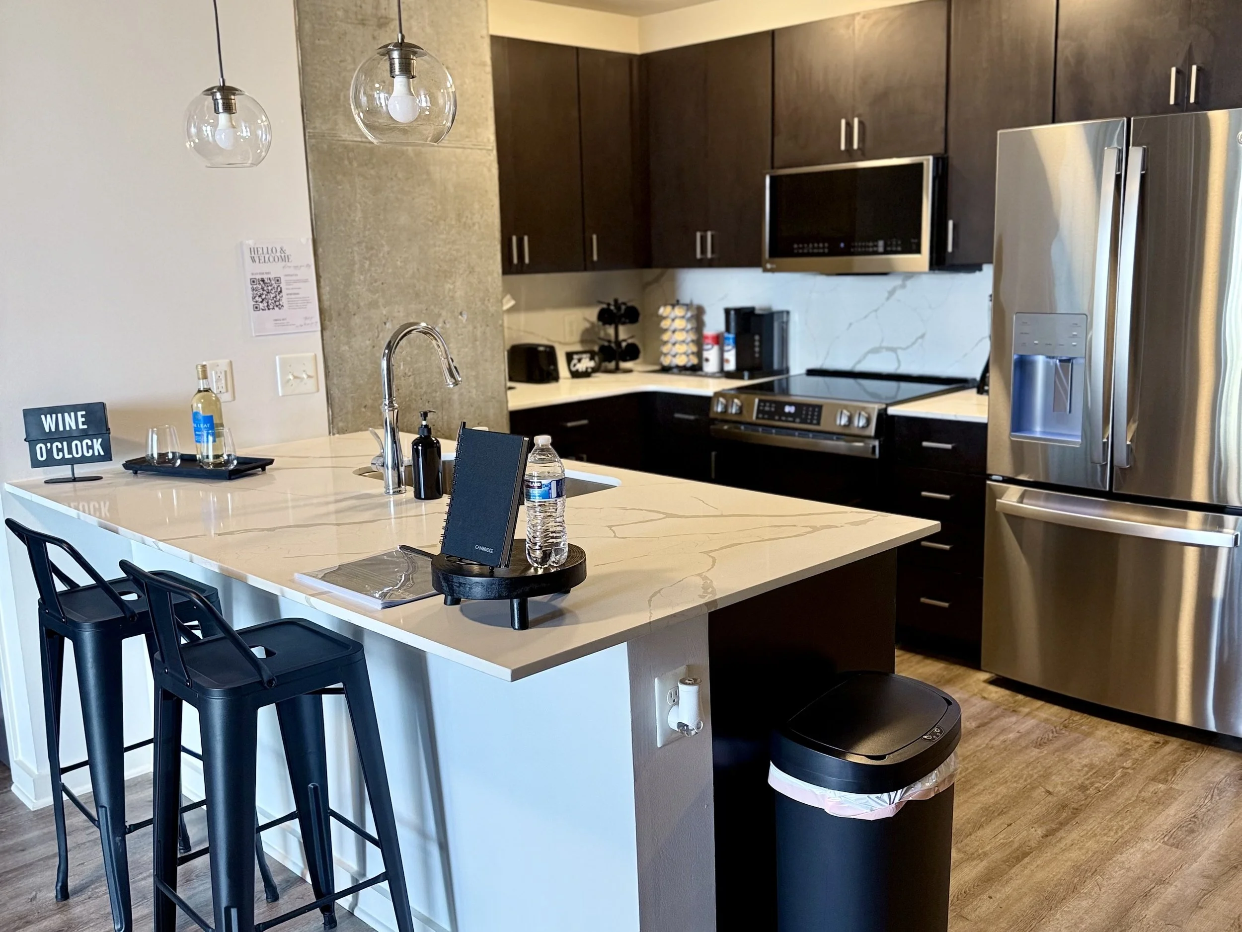Modern kitchen with white marble countertop island, dark brown cabinets, stainless steel appliances including a refrigerator and microwave, and hanging pendant lights. There are bar stools, a water bottle, a black sign that reads 'Wine O'Clock,' and 
