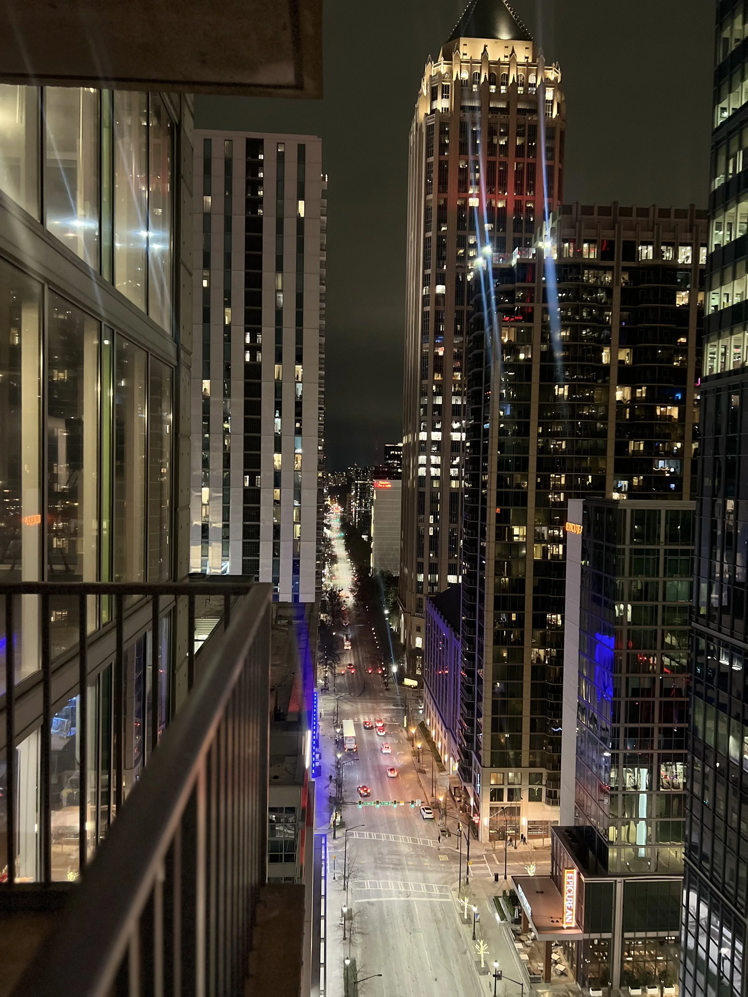 Night view of a city street lined with tall modern skyscrapers, some with illuminated windows, and a few lit signs, taken from a balcony with a metal railing in the foreground.