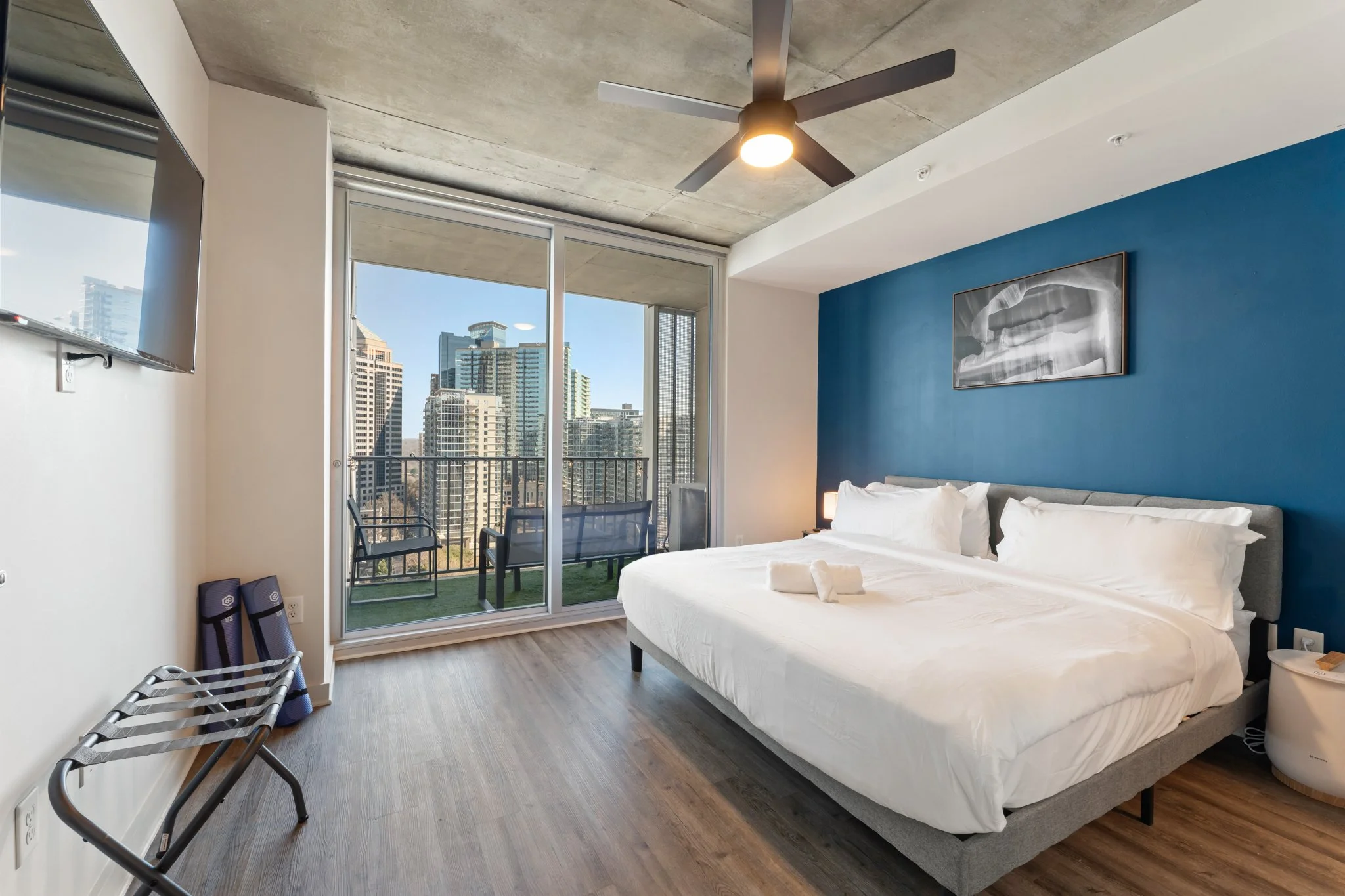 Modern hotel room with a large bed, blue accent wall, and a city view from a balcony, with a ceiling fan and artwork above the bed.